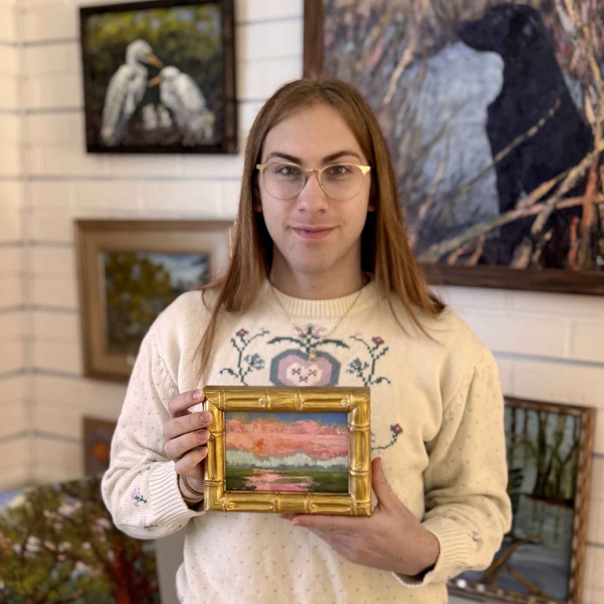 A Gray Man Gallery team member holding Blush of Day, a small framed original oil painting by Jeanne O’Neal featuring soft blush pink clouds, pale blue sky, and a quiet Lowcountry marsh landscape, shown in a gold bamboo-style frame and sized perfectly for bookshelf or easel display.