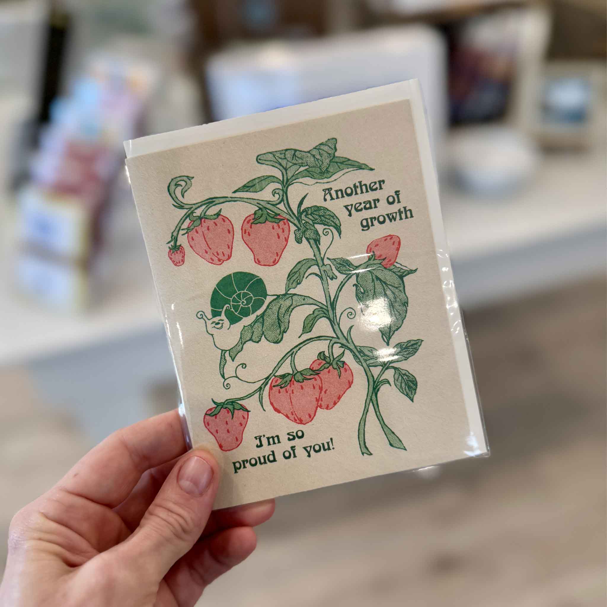 Another Year of Growth encouragement birthday card by Hannah-Michelle Bayley held inside Gray Man Gallery in Pawleys Island, featuring strawberries, leafy vines, a snail and the message “I’m so proud of you.”