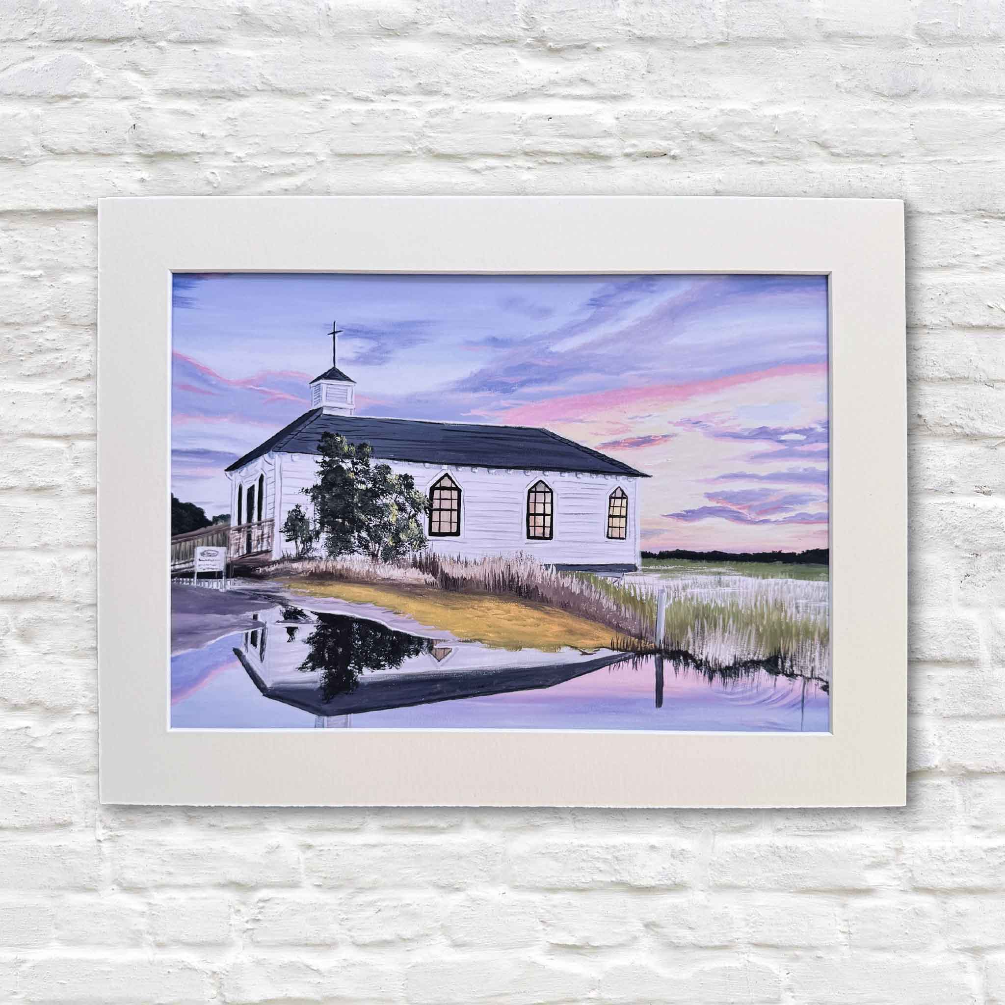 Pawleys Island Chapel fine art print by April Bensch displayed on a white wall, featuring a Lowcountry sunset scene with soft pink and blue skies, marsh reflections, and the iconic white chapel, matted to fit a standard 12 x 16 frame and available at Gray Man Gallery in Pawleys Island.
