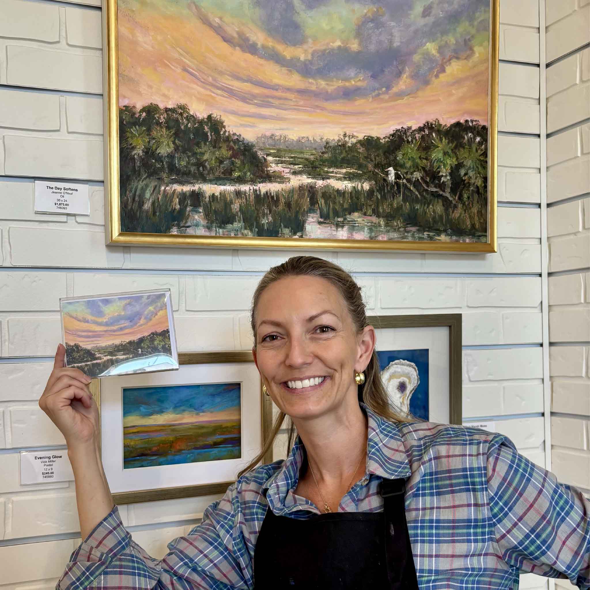Artist Jeanne O'Neal holds the matching The Day Softens notecard beneath her original oil painting at Gray Man Gallery in Pawleys Island, showcasing the collectible card alongside the original Lowcountry marsh landscape.