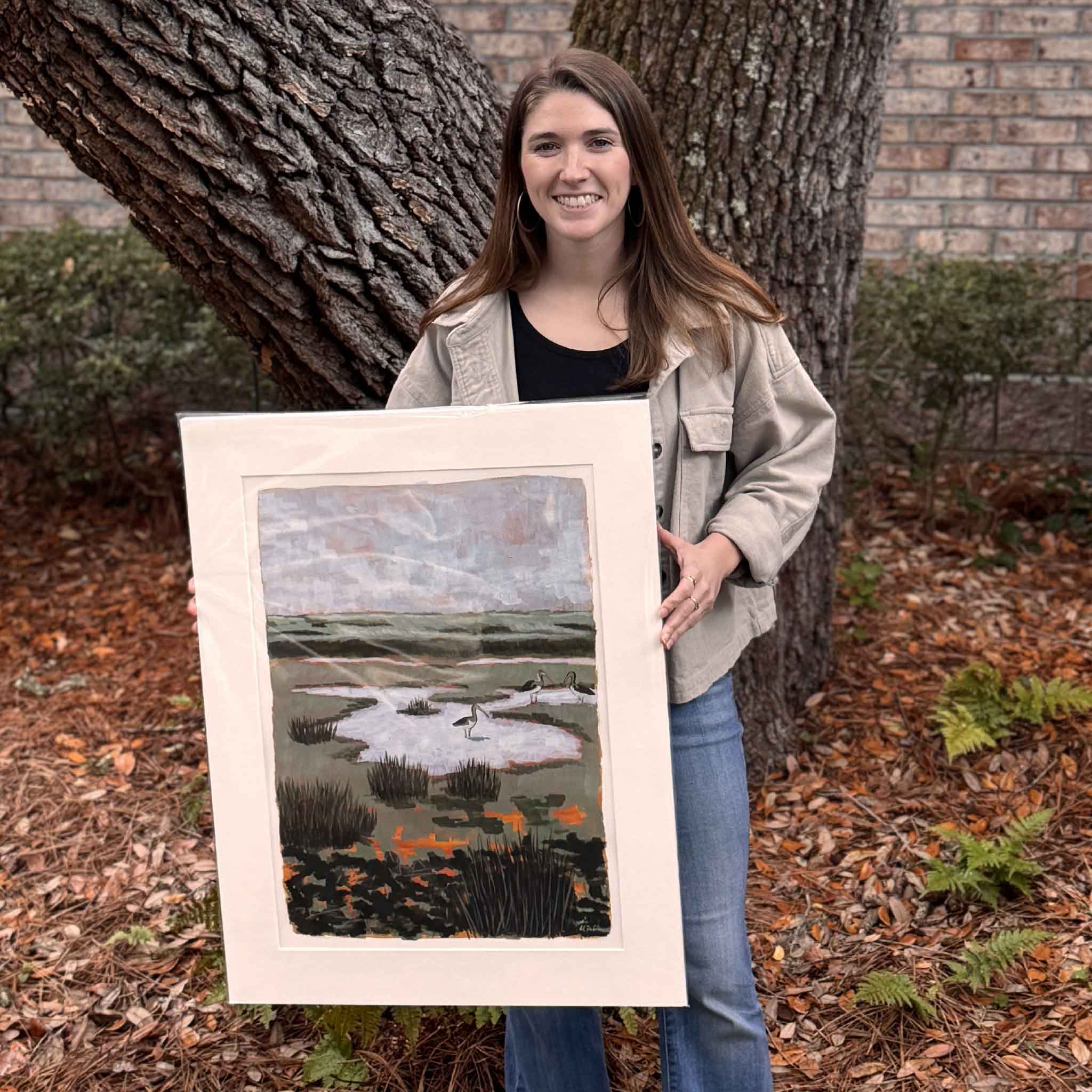 Local artist Maggie Pelton holding her original acrylic painting Ibis at North Inlet, matted to 22 x 28 inches, shown for scale inside Gray Man Gallery in Pawleys Island, South Carolina.