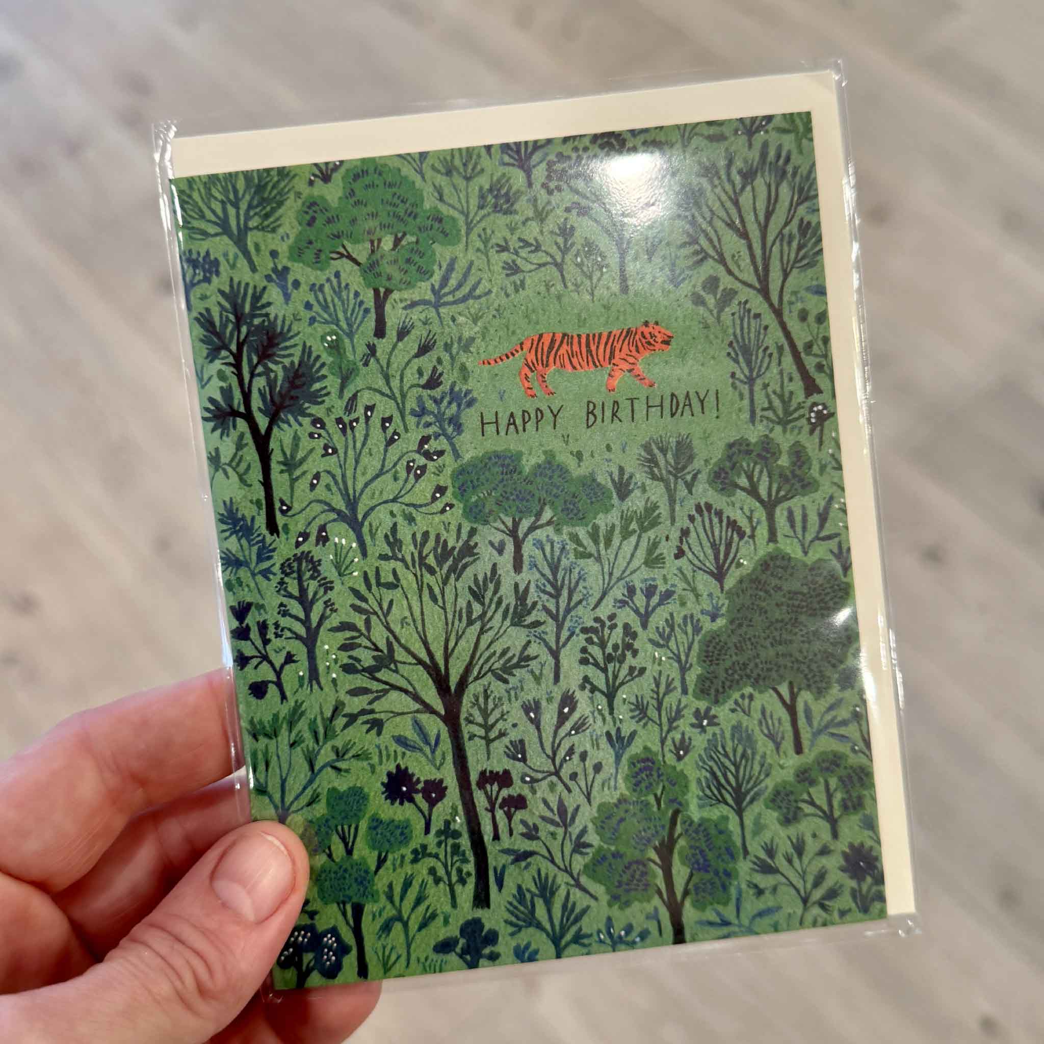 Orange Tiger Birthday greeting card by Becca Stadtlander from Red Cap Cards, photographed inside Gray Man Gallery in Pawleys Island, South Carolina. This premium birthday card features a vibrant orange tiger walking through an intricate green forest landscape and is printed on heavyweight uncoated cardstock with a matching envelope.