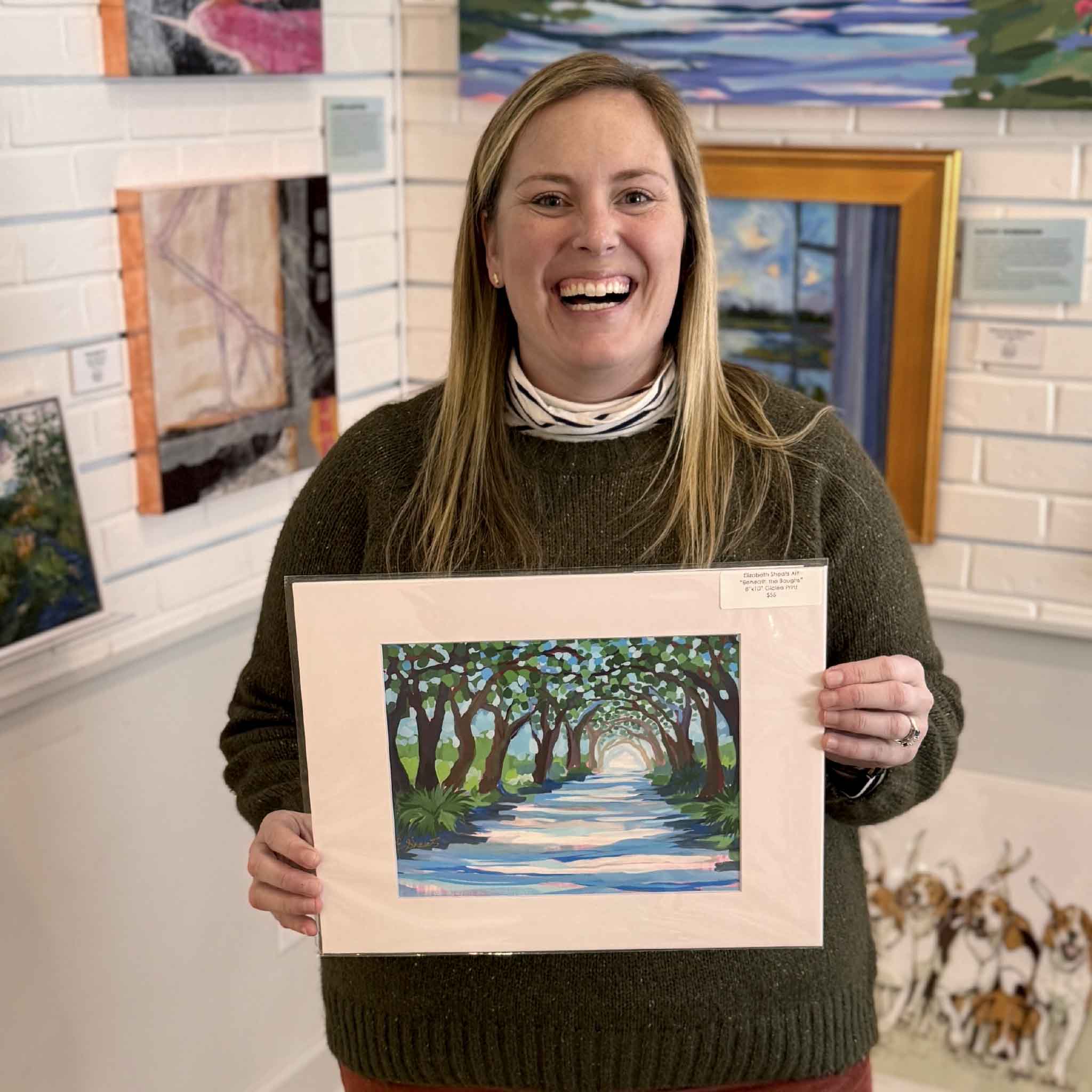 Bordy holding Elizabeth Sheats Beneath the Boughs 11x14 fine art giclée print at Gray Man Gallery in Pawleys Island South Carolina, featuring live oak tree canopy, shaded plantation road, and peaceful Lowcountry landscape artwork by local artist