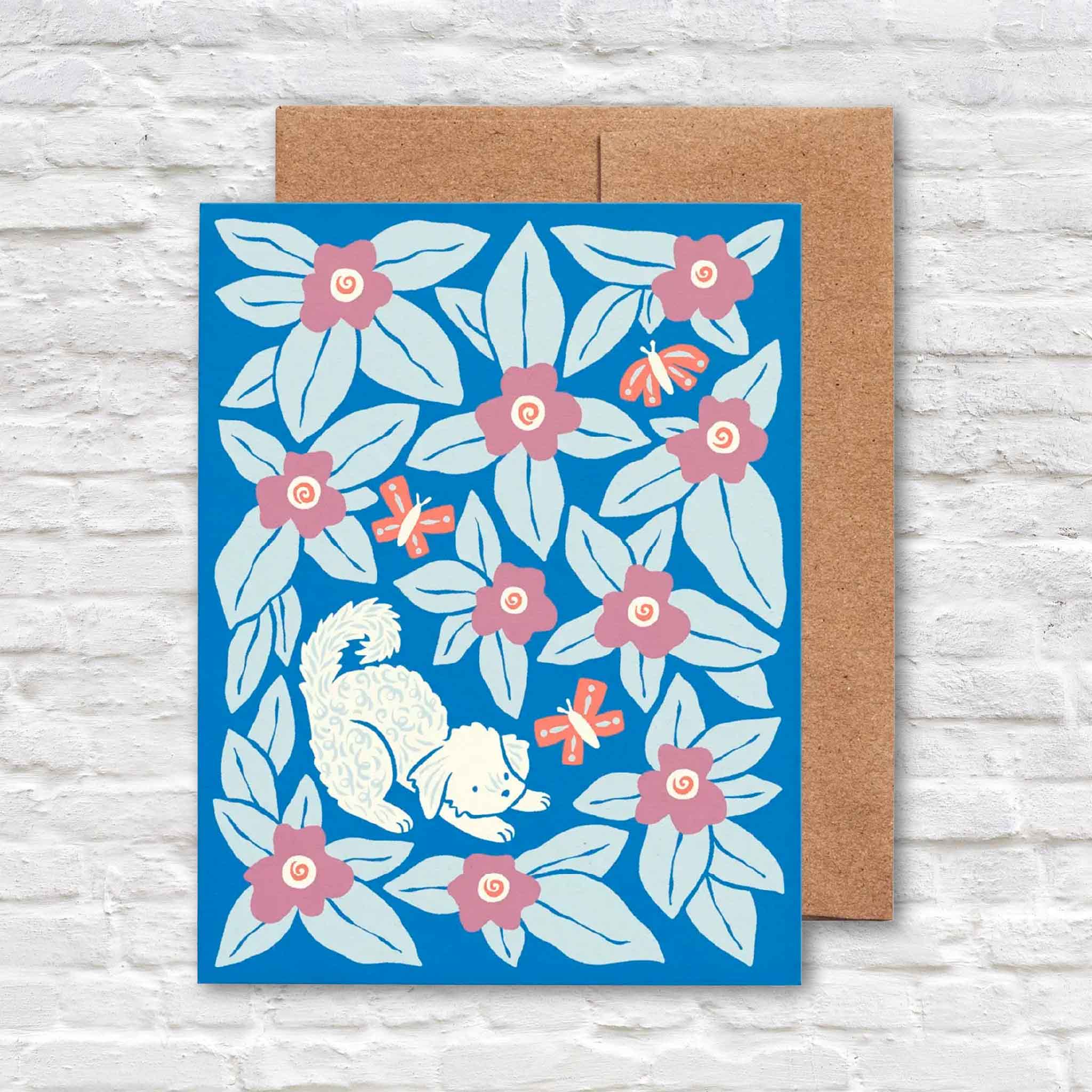 Bennie Blooms greeting card by Chrissie Van Hoever featuring a playful curly-coated white dog chasing butterflies among pink flowers and blue foliage with a recycled kraft envelope. Available at Gray Man Gallery in Pawleys Island, South Carolina.