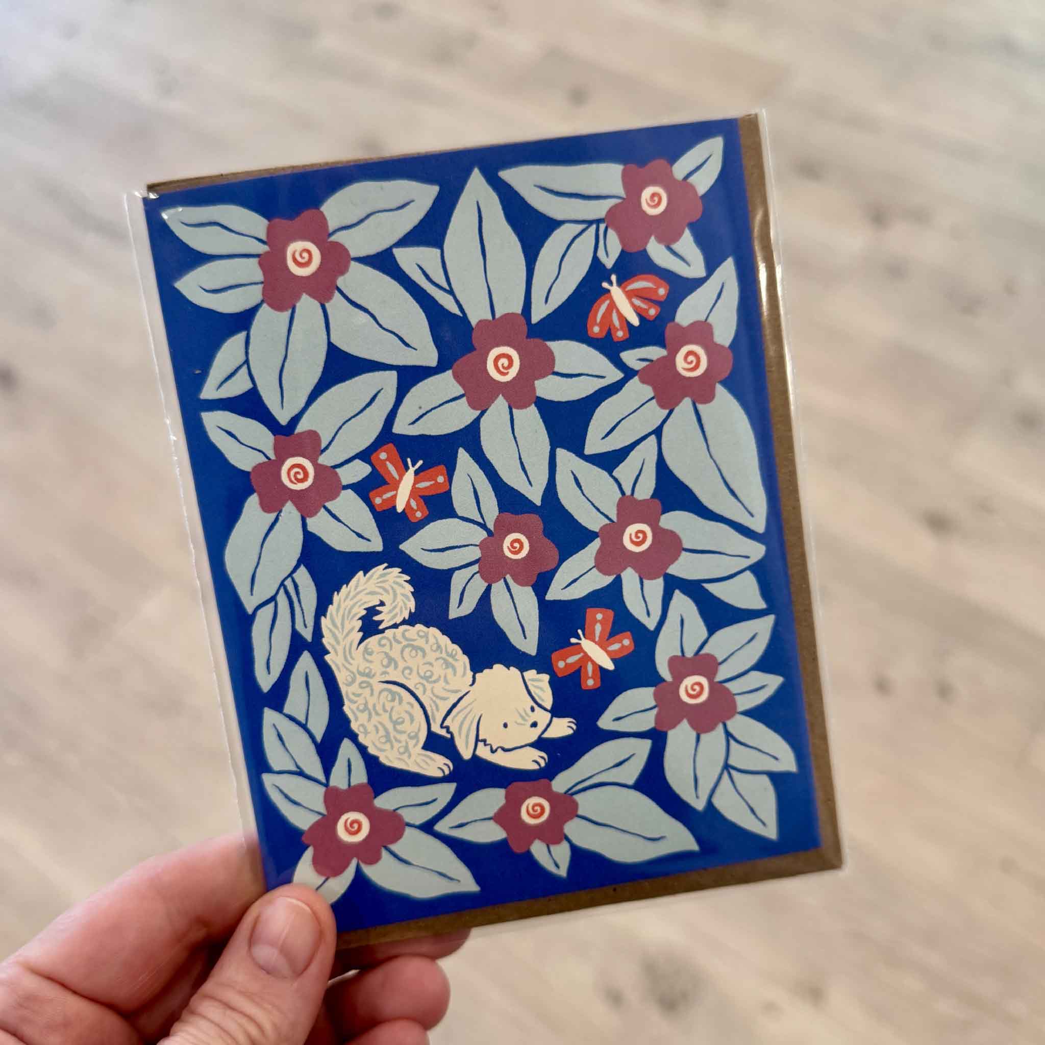 Customer holding the Bennie Blooms Greeting Card by Chrissie Van Hoever inside Gray Man Gallery in Pawleys Island, South Carolina. This whimsical Nuthatch Studio greeting card features Bennie, a playful curly-coated dog surrounded by bright blue flowers and butterflies, printed on premium 100% recycled paper and paired with a recycled kraft envelope.