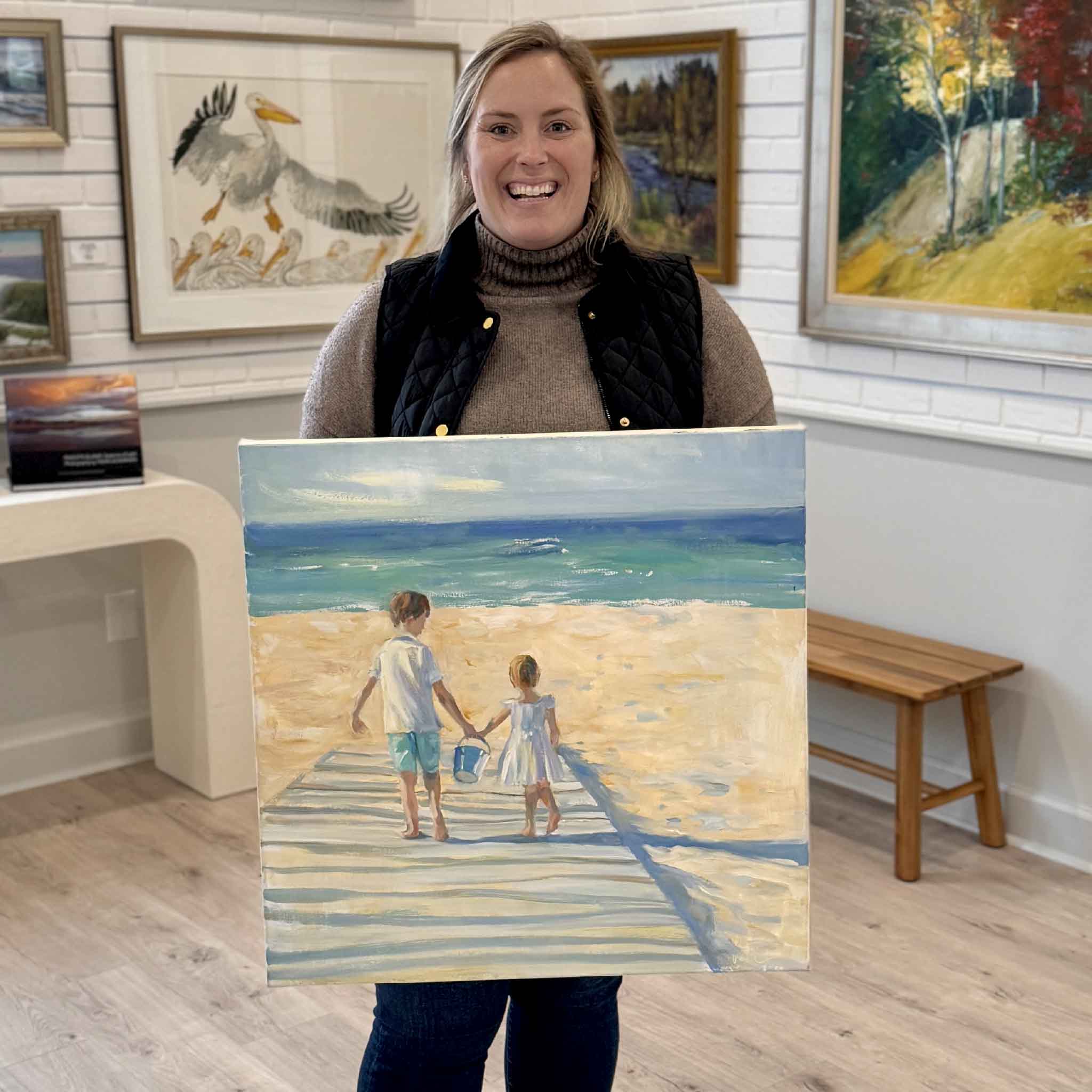 Bordy holding “Best Friends,” an original oil painting by Helli Luck at Gray Man Gallery in Pawleys Island, featuring two children walking hand in hand on a beach boardwalk with turquoise ocean waves, showcasing local Lowcountry coastal art and family-inspired storytelling
