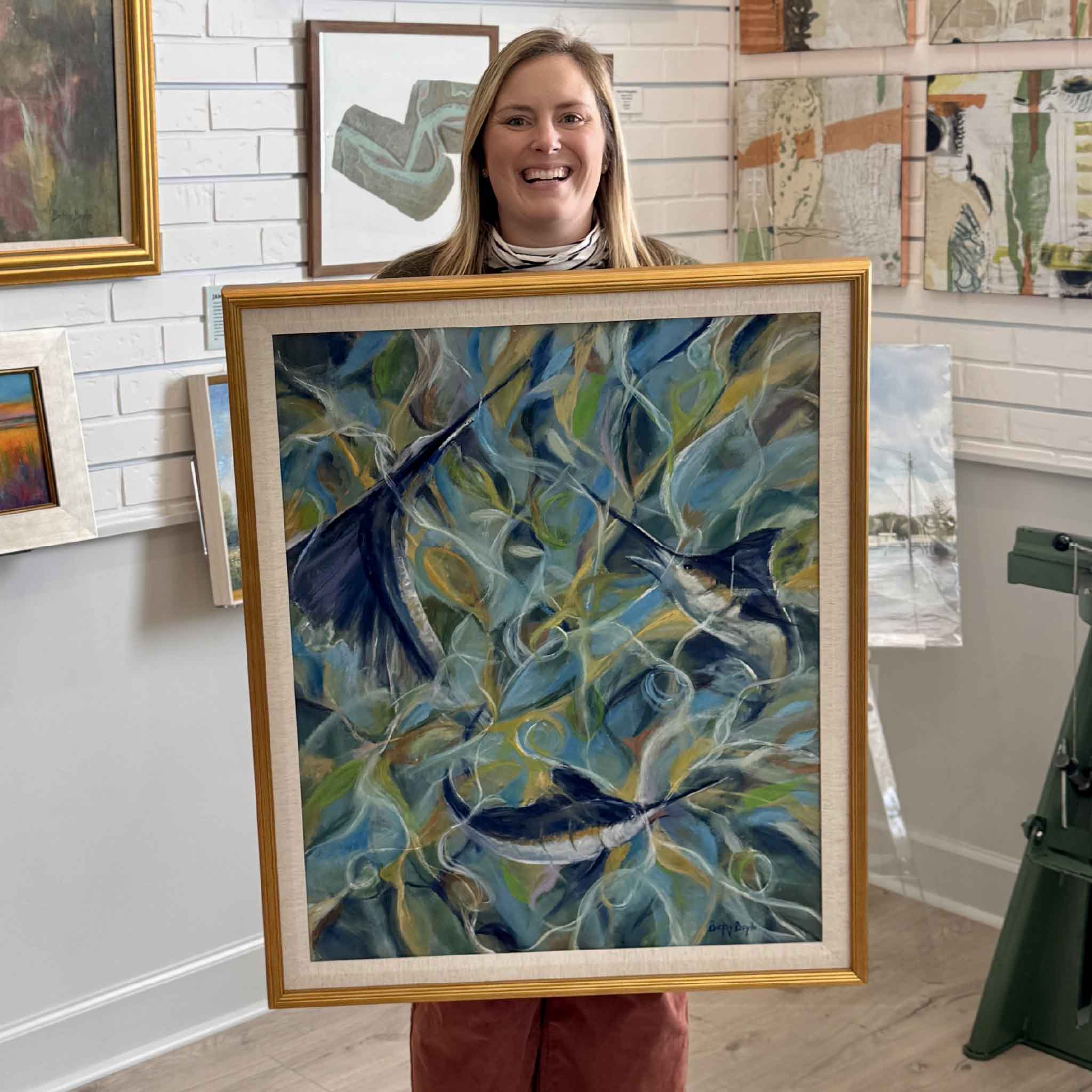 Bordy holding Betsy Boyle’s “Circling” original oil painting at Gray Man Gallery in Pawleys Island, showing scale of framed coastal artwork by local South Carolina artist featuring billfish in motion