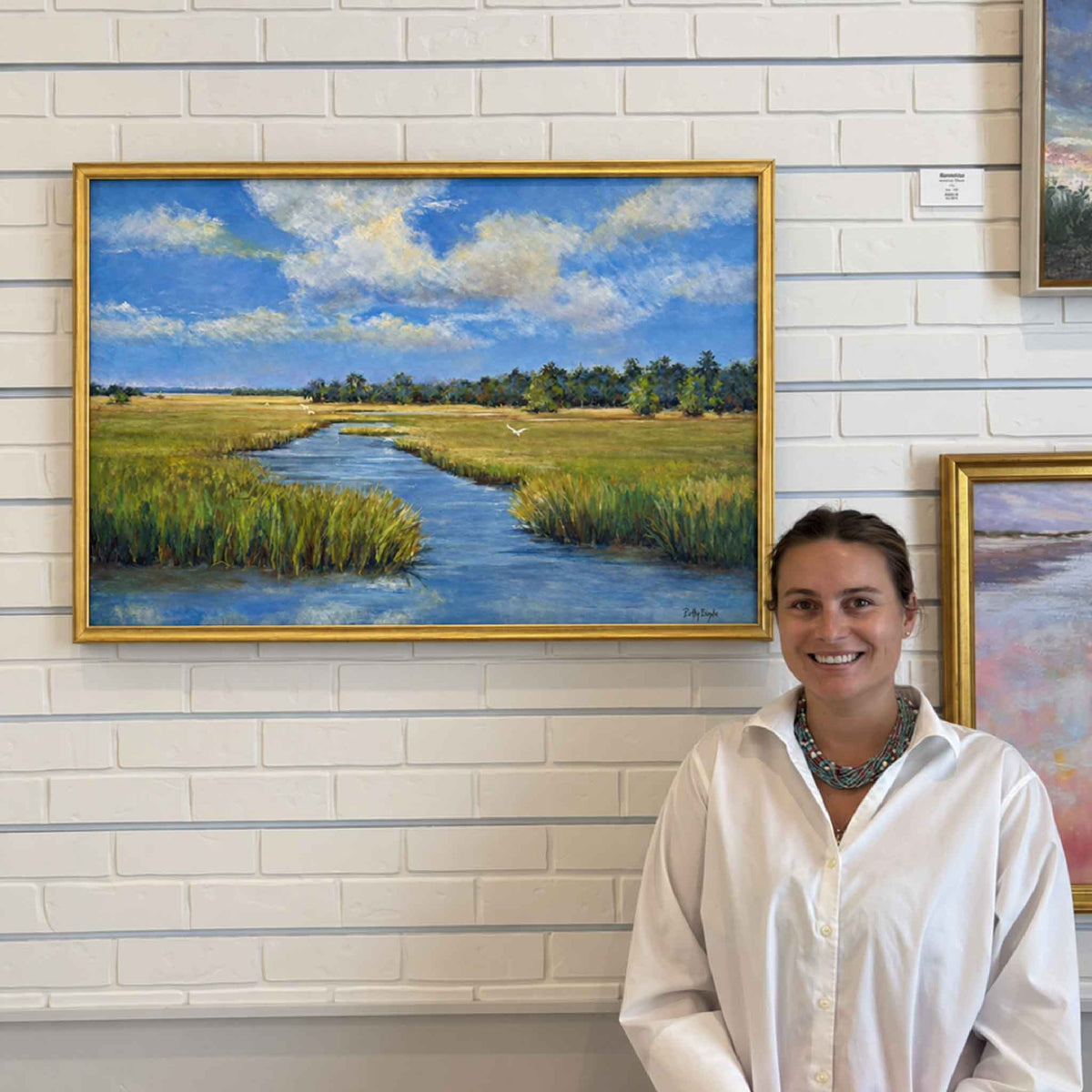 Marsh Life by local artist Betsy Boyle displayed at Gray Man Gallery in Pawleys Island, South Carolina, showing an original framed oil painting of a blue tidal creek winding through green Lowcountry marsh beneath a bright cloud-filled coastal sky