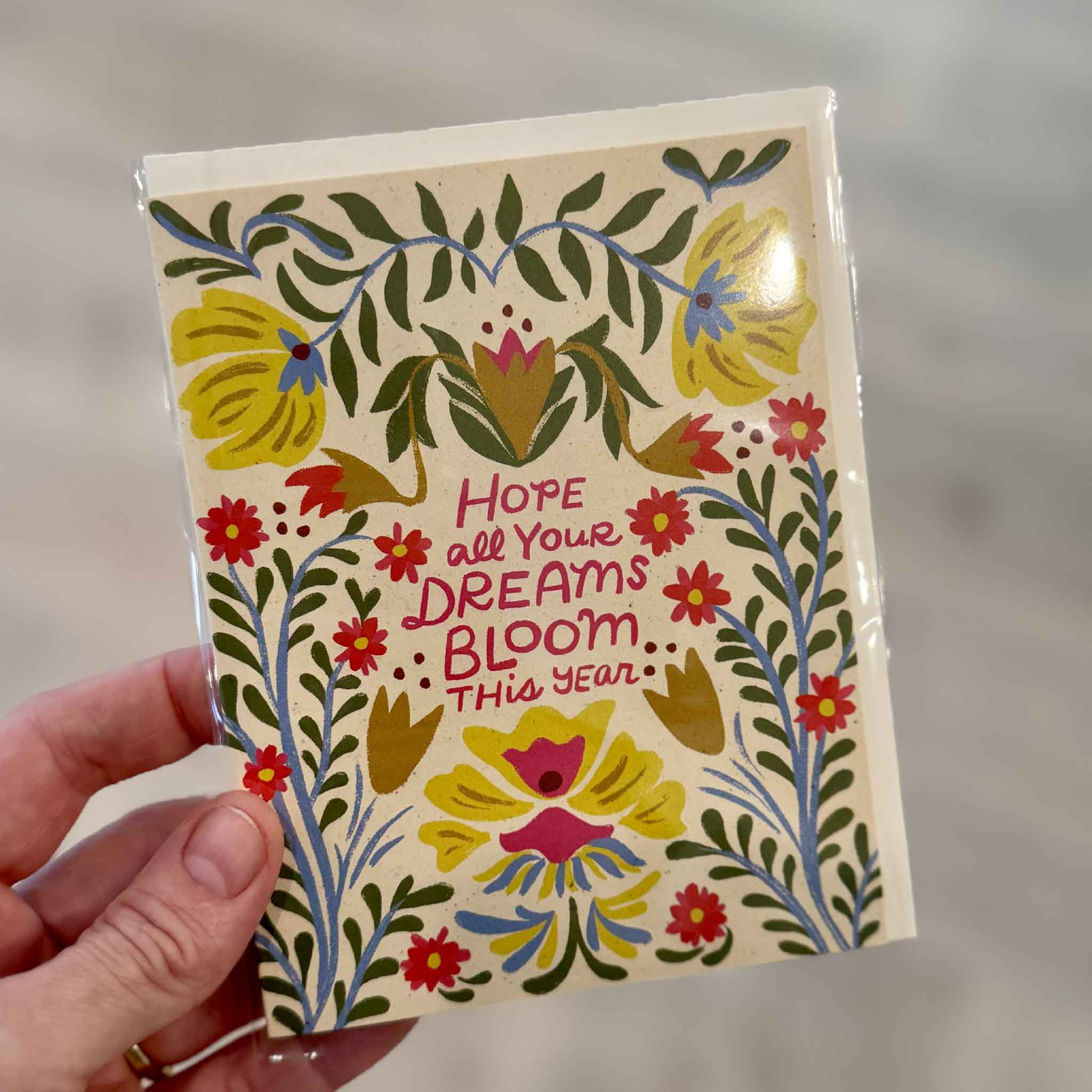 A hand holds the Blooming Dreams greeting card by Dylan Mierzwinski inside Gray Man Gallery in Pawleys Island, South Carolina. Featuring vibrant folk-inspired florals, lush greenery, and the inspiring message "Hope all your dreams bloom this year," this blank-inside greeting card is a thoughtful choice for birthdays, graduations, encouragement, new jobs, retirements, and celebrating life's exciting new chapters.