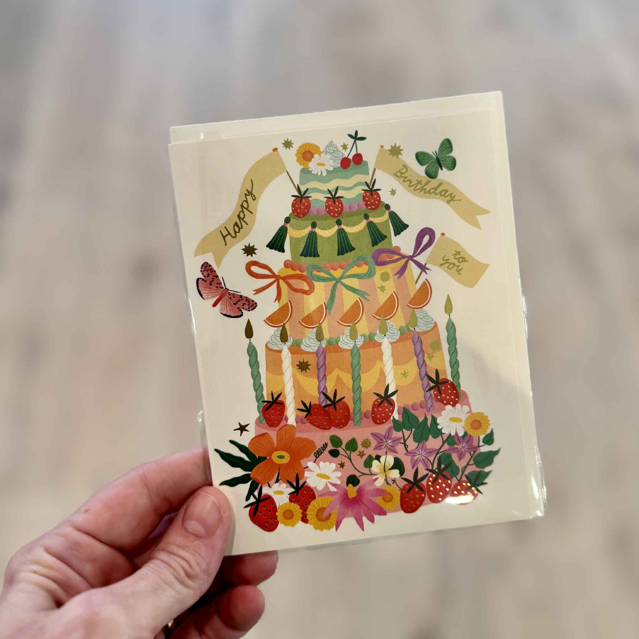 Whimsical Cake birthday greeting card by Bodil Jane shown inside Gray Man Gallery in Pawleys Island, featuring a bright maximalist tiered cake with flowers, strawberries, ribbons, candles, butterflies and festive gold foil accents.