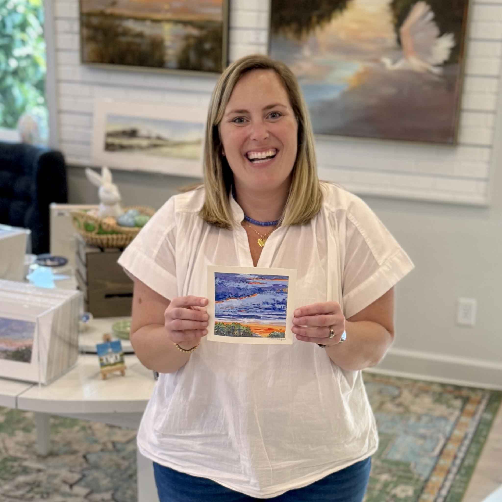 Bordy holding Afterglow by Jeanne O’Neal inside Gray Man Gallery in Pawleys Island showing the scale of the 6x6 original gouache sunset with dramatic violet clouds and bright orange horizon