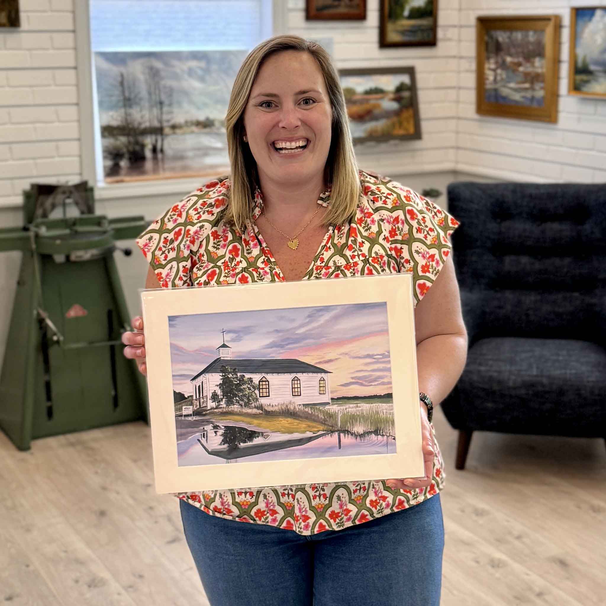 Bordy holding a signed Pawleys Island Chapel fine art print by April Bensch inside Gray Man Gallery, showing the scale of the piece, a serene Lowcountry sunset with marsh reflections and pastel skies, professionally matted to fit a standard 12 x 16 frame for easy framing.