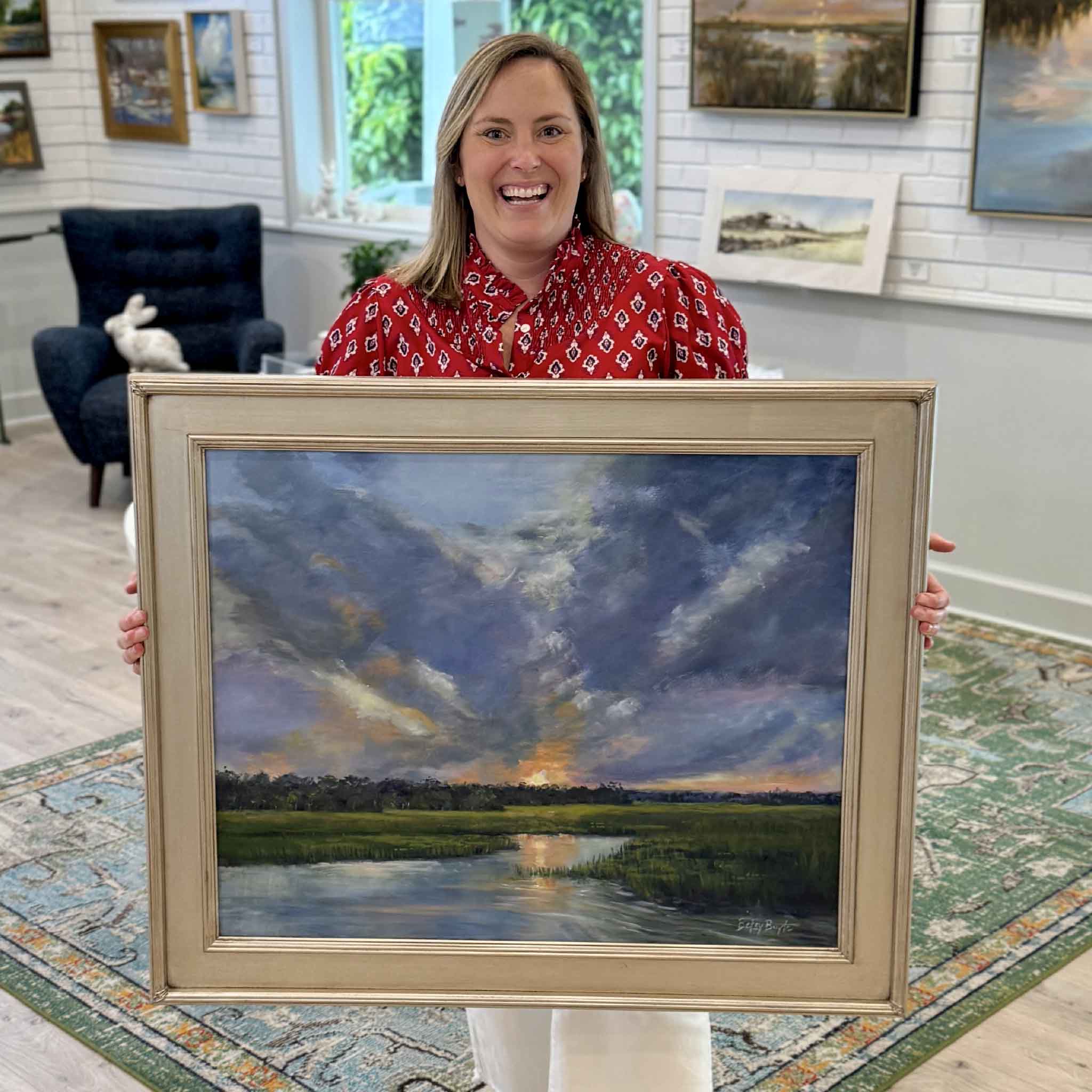 Bordy holding Dramatic Exit by Betsy Boyle inside Gray Man Gallery in Pawleys Island, South Carolina, showing the scale of the framed original oil painting with a Lowcountry marsh sunset, expansive cloud-filled sky, and reflective water illuminated by warm horizon light