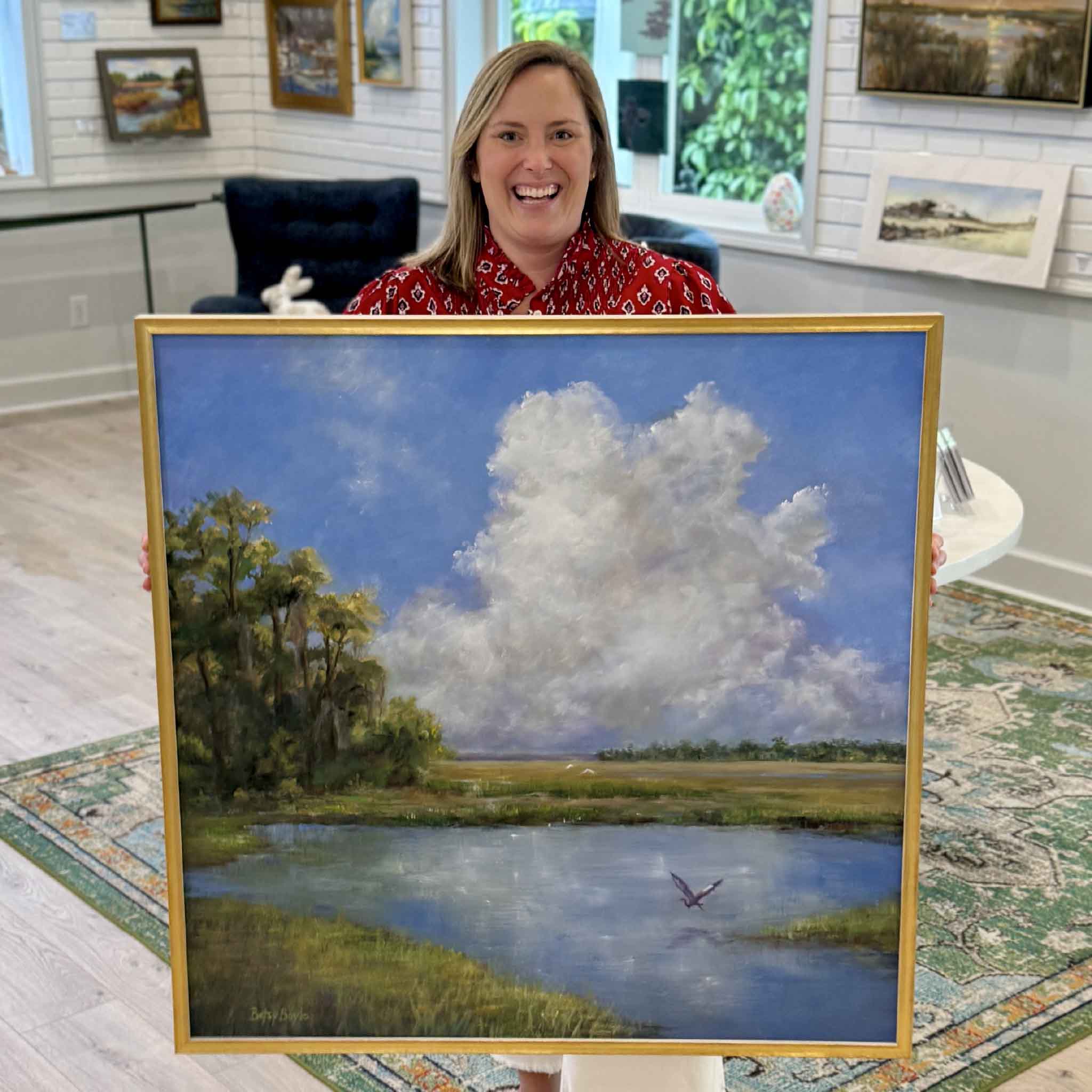 Bordy holding Just Breathe by Betsy Boyle inside Gray Man Gallery in Pawleys Island, South Carolina, showing the scale of the framed original oil painting with a Lowcountry marsh scene, expansive sky filled with soft clouds, reflective water, and coastal landscape details