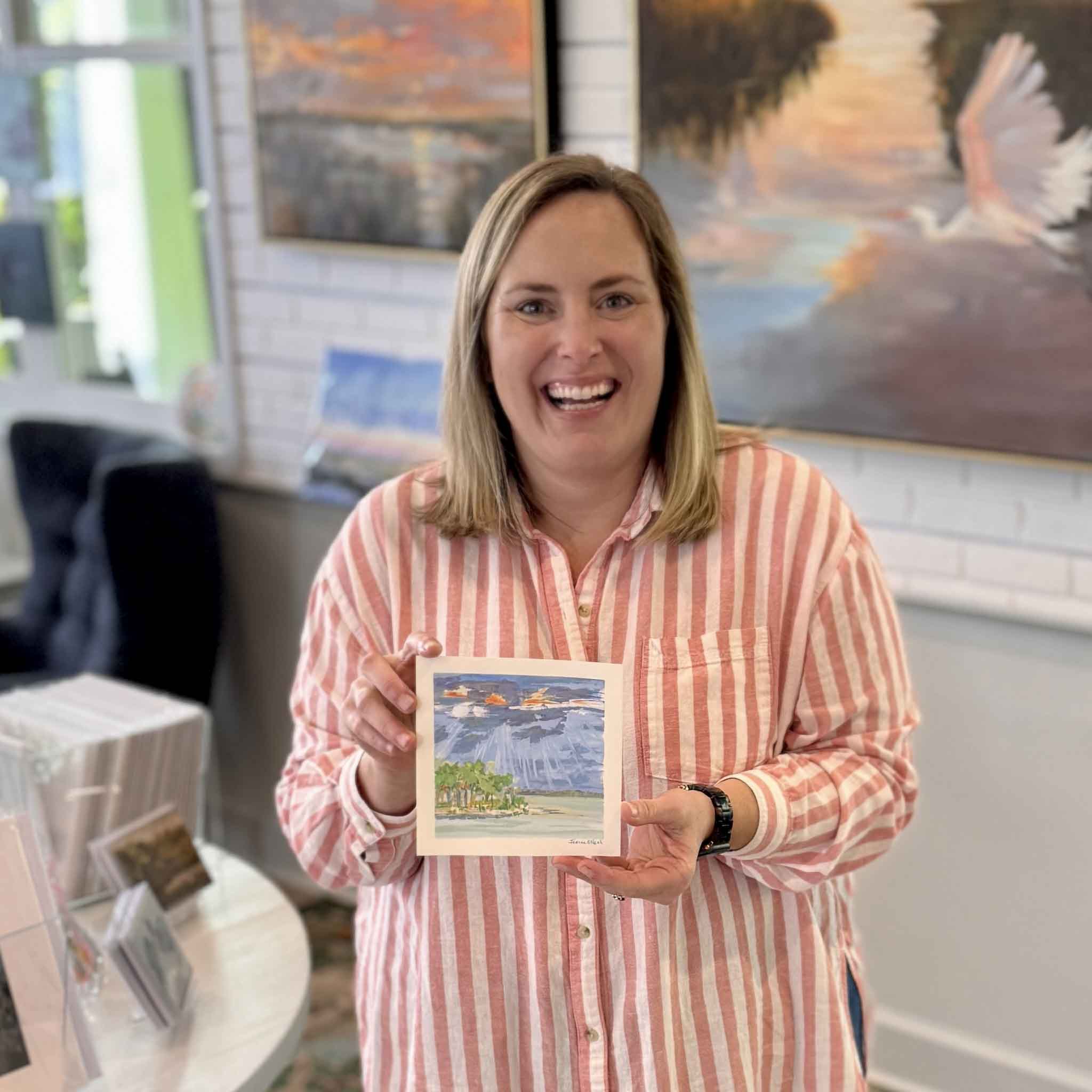 Bordy holding Breaking Light by Jeanne O’Neal inside Gray Man Gallery in Pawleys Island showing the scale of the 6x6 original gouache coastal landscape with bold sun rays through stormy clouds over calm water