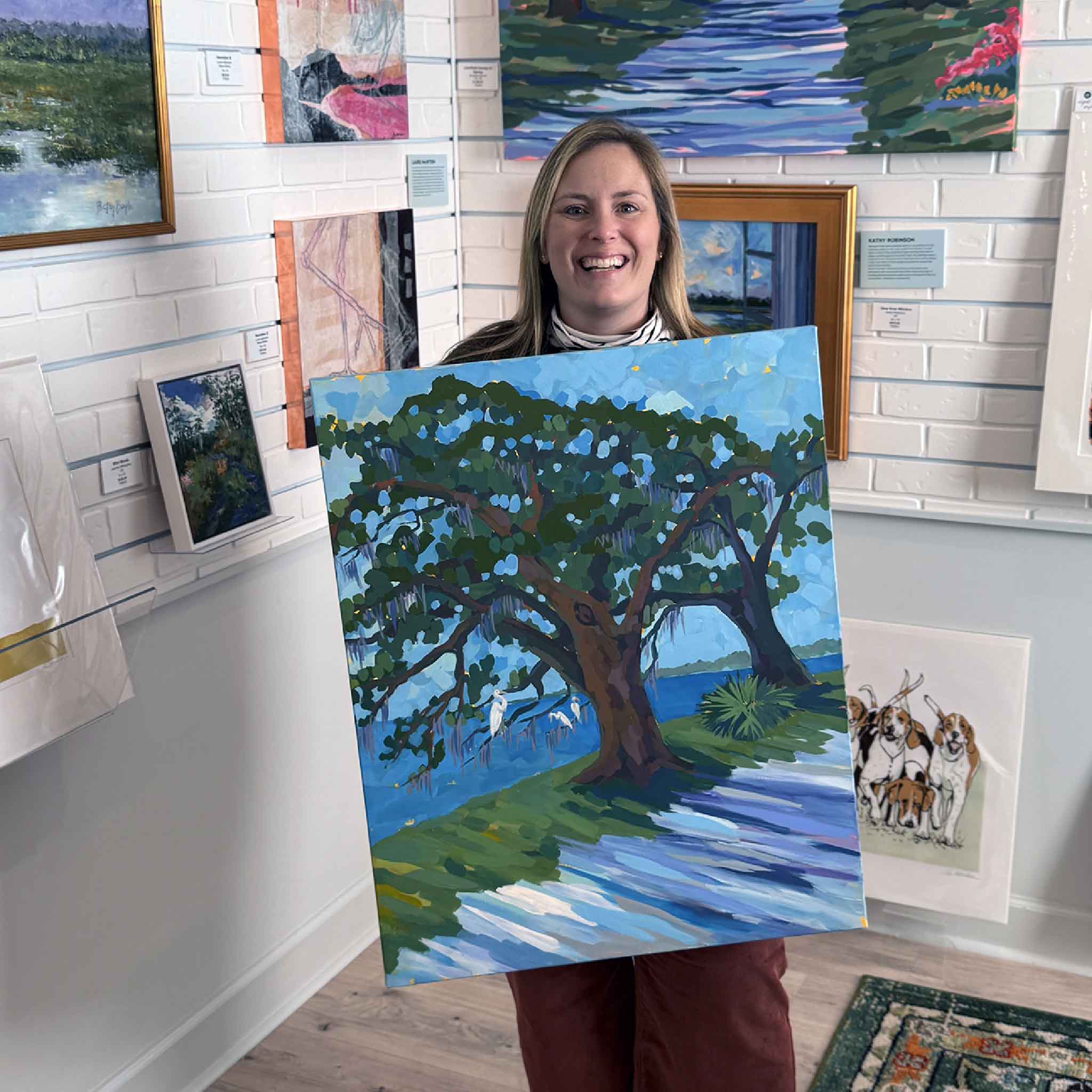 Gray Man Gallery team member Bordy holding the original painting Canopy Walk I by Elizabeth Sheats inside the Pawleys Island gallery, showcasing local Lowcountry artwork