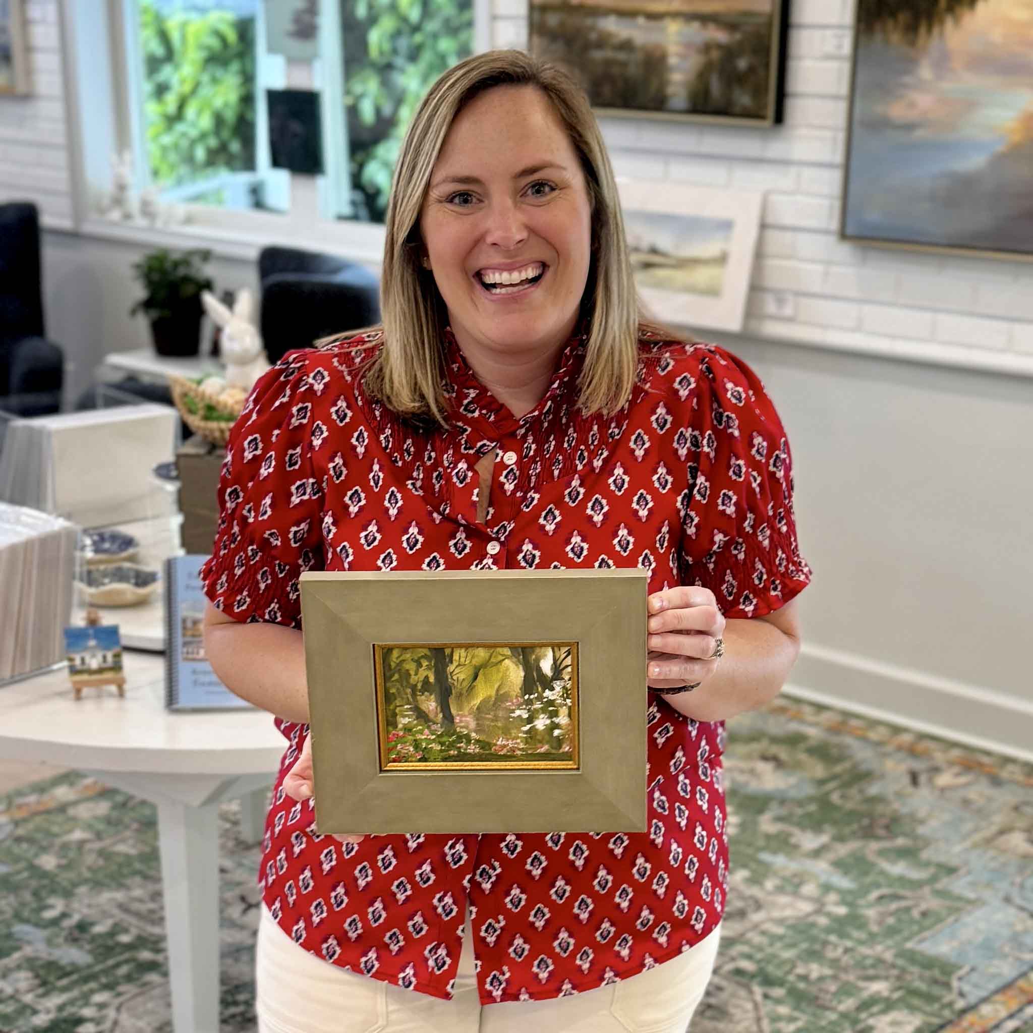 Bordy holding Evening Garden by Jeanne O’Neal original oil painting created at Brookgreen Gardens inside Gray Man Gallery Pawleys Island showing scale of framed floral garden landscape with warm evening light