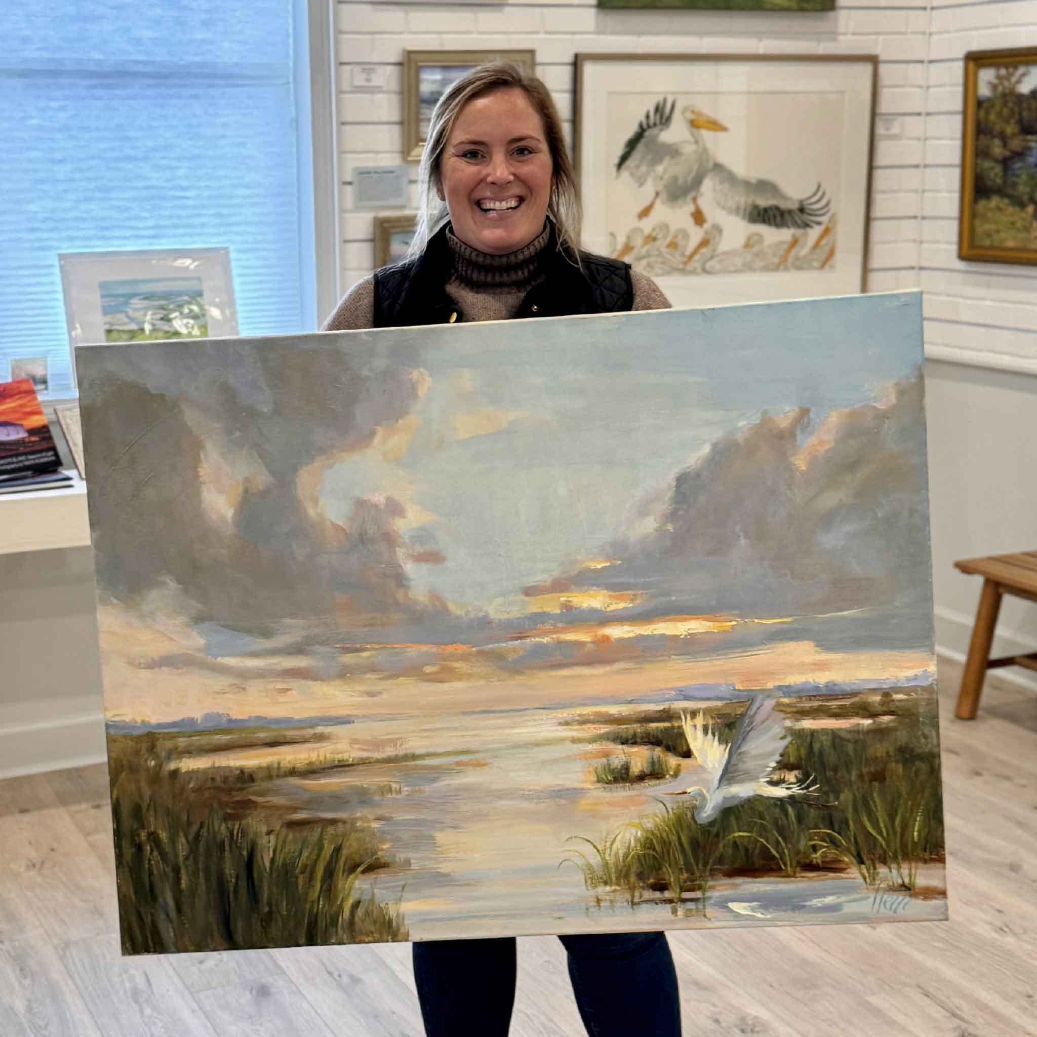 Gray Man Gallery staff member Bordy holding “Rising with the Dawn,” a large original oil painting by local Lowcountry artist Helli Luck, inside Gray Man Gallery in Pawleys Island, South Carolina, featuring coastal marsh landscape artwork, wildlife imagery, and expert custom framing services