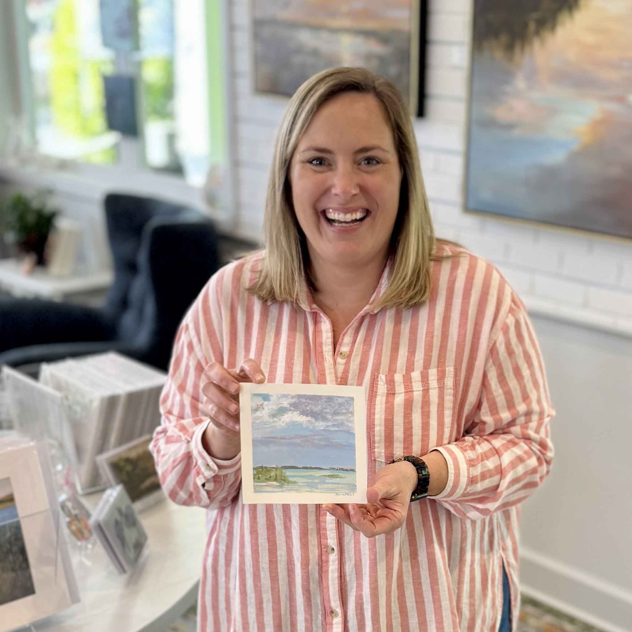 Bordy holding At Anchor by Jeanne O’Neal inside Gray Man Gallery in Pawleys Island, showing the scale of the 6x6 original gouache painting with a small boat at rest in pale blue and green Abaco waters beneath a soft layered sky