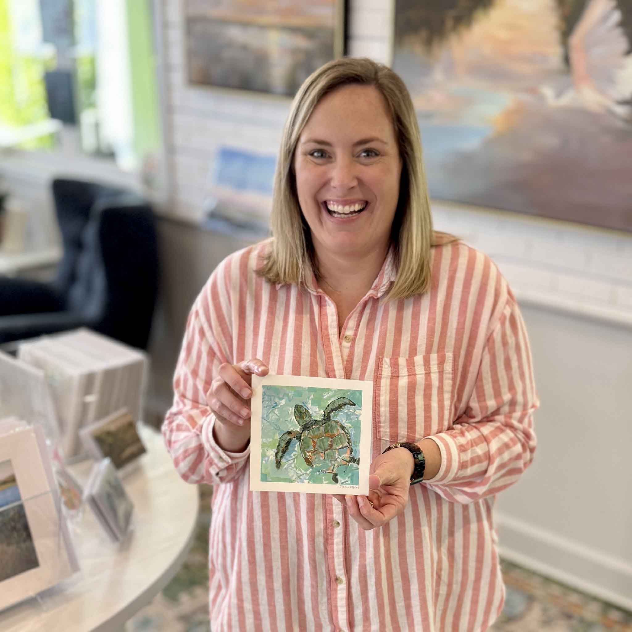 Bordy holding Drift Current by Jeanne O’Neal inside Gray Man Gallery in Pawleys Island, 6x6 original gouache painting of a sea turtle in motion with layered coastal colors and textured brushwork