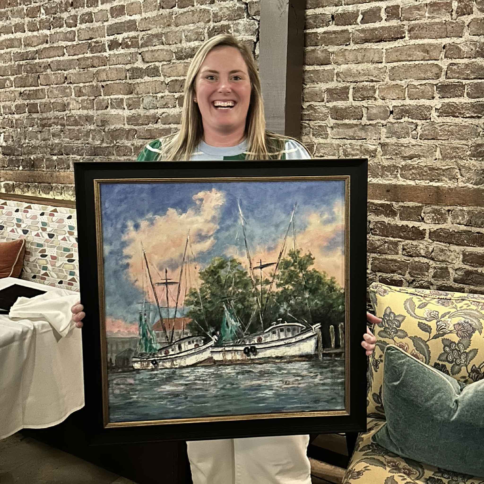 Bordy of Gray Man Gallery holding the original oil painting “End of Day” by Jeanne O’Neal while standing inside 631 Frank's on Front Restaurant in Georgetown, South Carolina, where the artwork is currently on display, showcasing the professionally custom framed coastal scene of shrimp boats at sunset and highlighting the scale, detail, and craftsmanship of this Lowcountry maritime artwork presented by Pawleys Island’s trusted local art gallery and custom frame shop