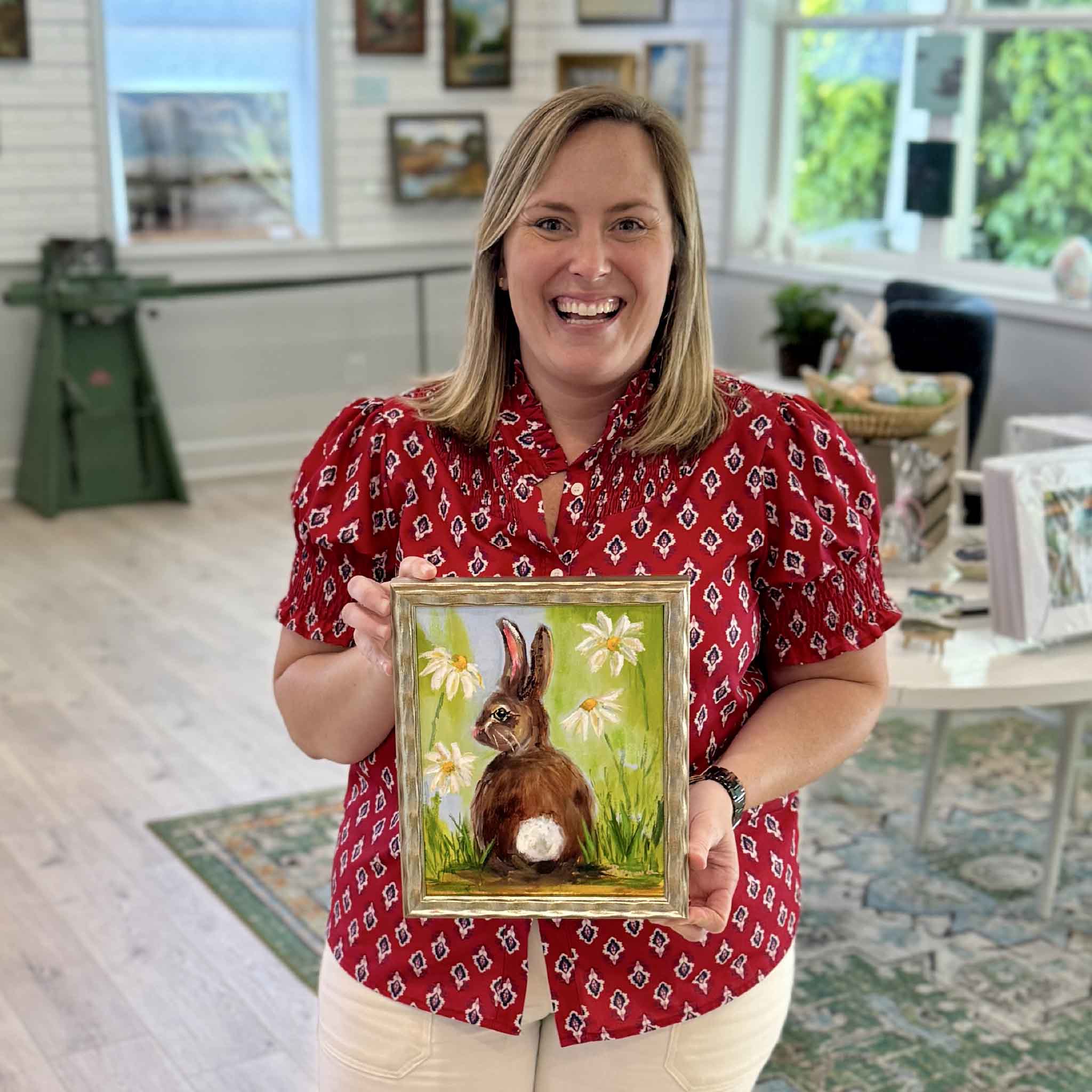 Bordy holding Spring Forward by Jenny Reynolds inside Gray Man Gallery in Pawleys Island, South Carolina, showing the scale of the framed original oil painting of a bunny surrounded by daisies in a bright spring landscape with cheerful colors and painterly brushwork