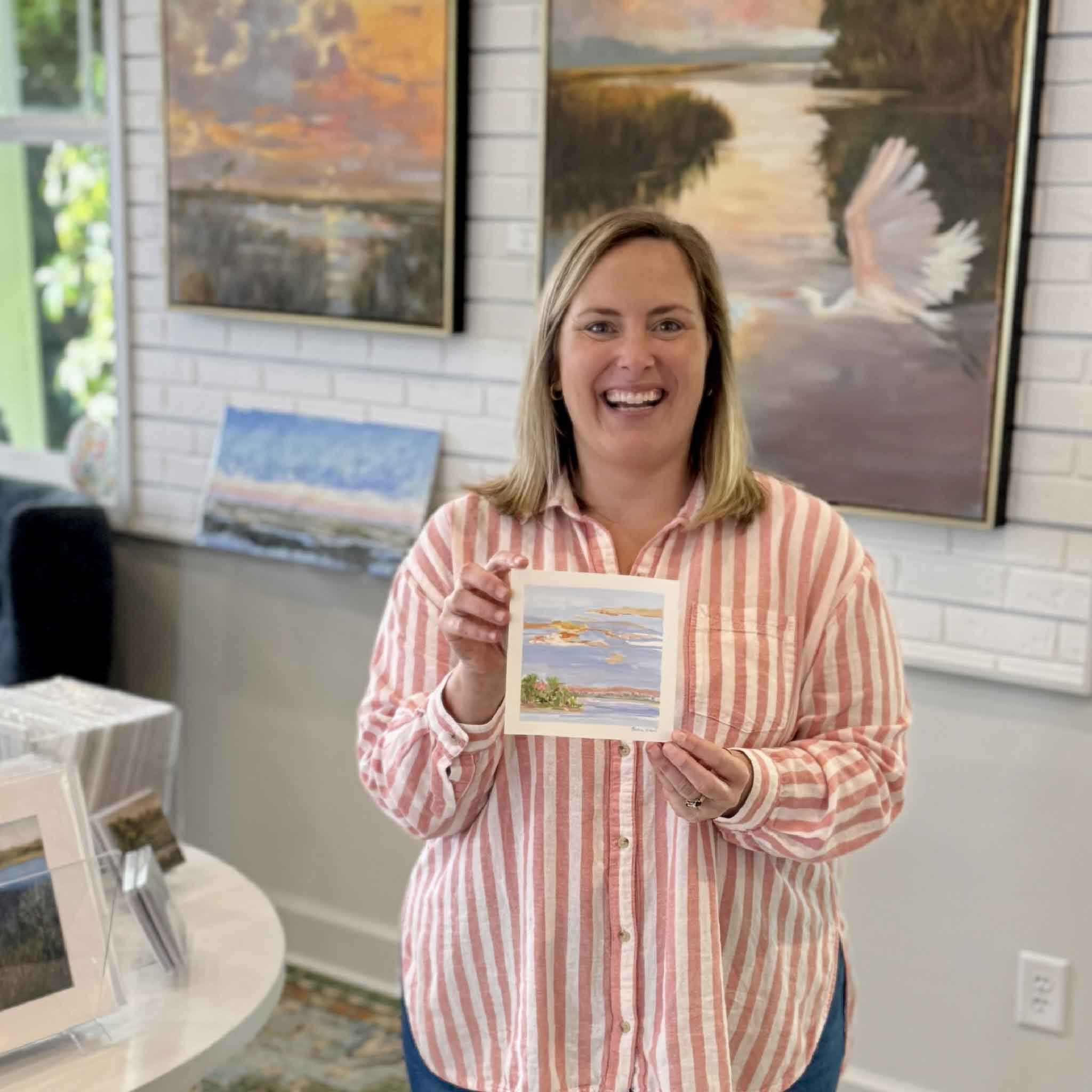 Bordy holding Last Light Over Abaco by Jeanne O’Neal inside Gray Man Gallery in Pawleys Island showing the scale of the 6x6 original gouache coastal landscape with warm sunset sky and calm blue water from the Postcards from Abaco collection