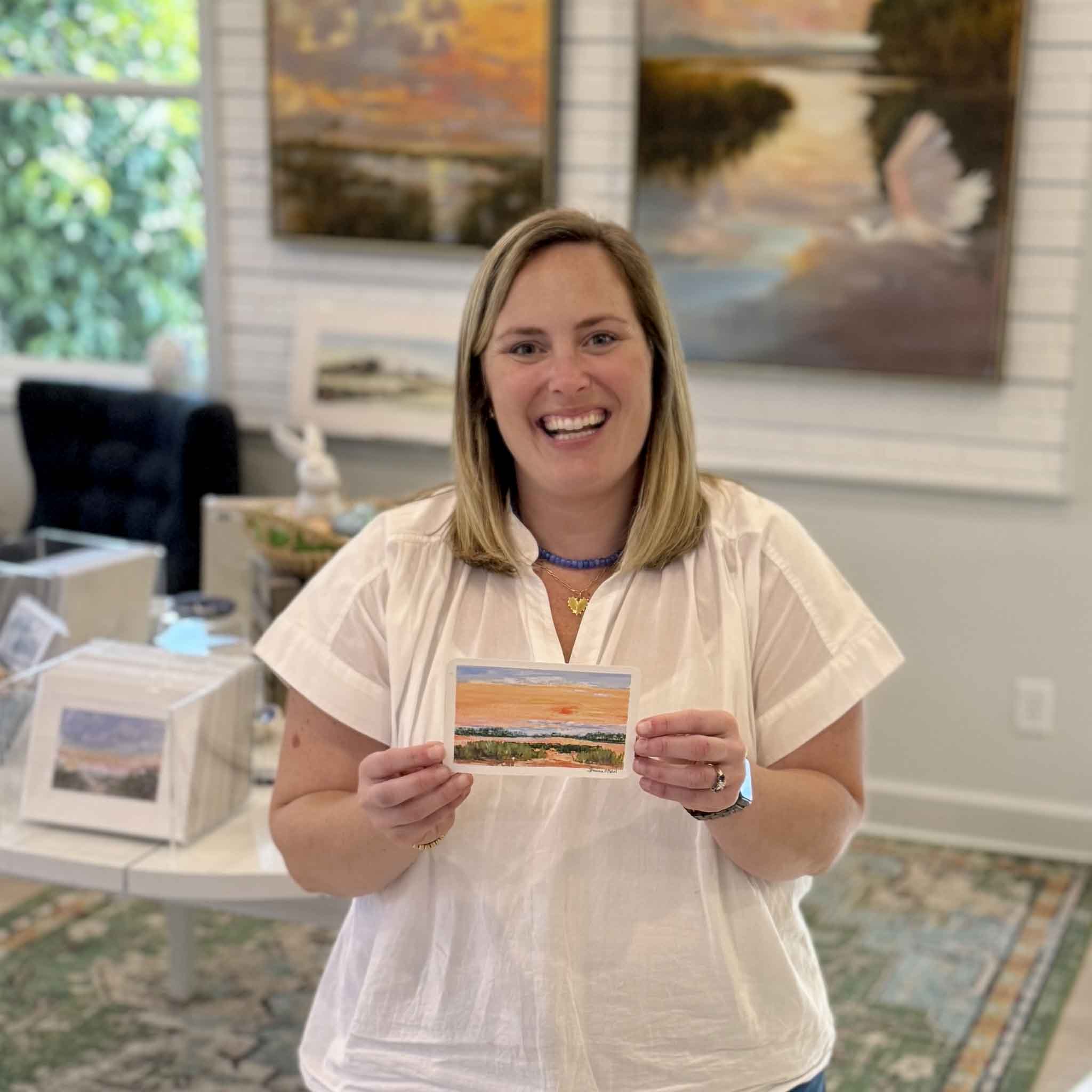 Bordy holding Low Sun by Jeanne O’Neal inside Gray Man Gallery in Pawleys Island showing the scale of the 6x4 original gouache sunset painted on postcard with hazy orange sky and low sun over coastal landscape
