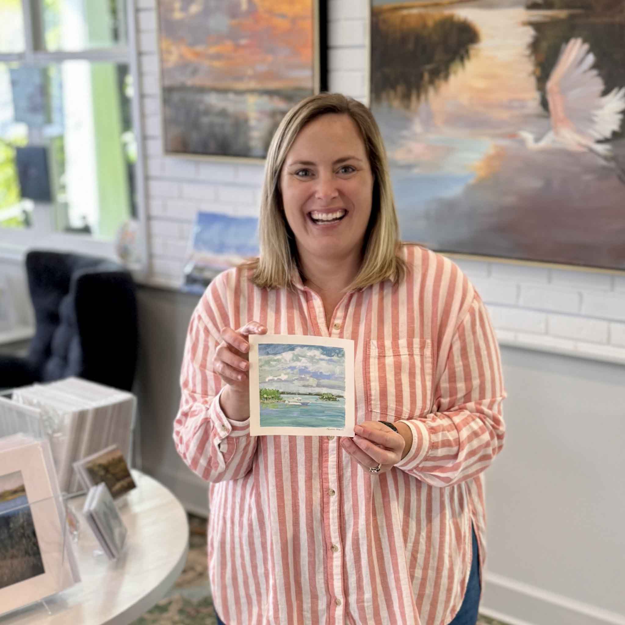 Bordy holding Open Water by Jeanne O’Neal inside Gray Man Gallery in Pawleys Island showing the scale of the 6x6 original gouache coastal landscape with expansive blue sky, layered clouds, shallow turquoise water, and distant low islands from the Postcards from Abaco collection