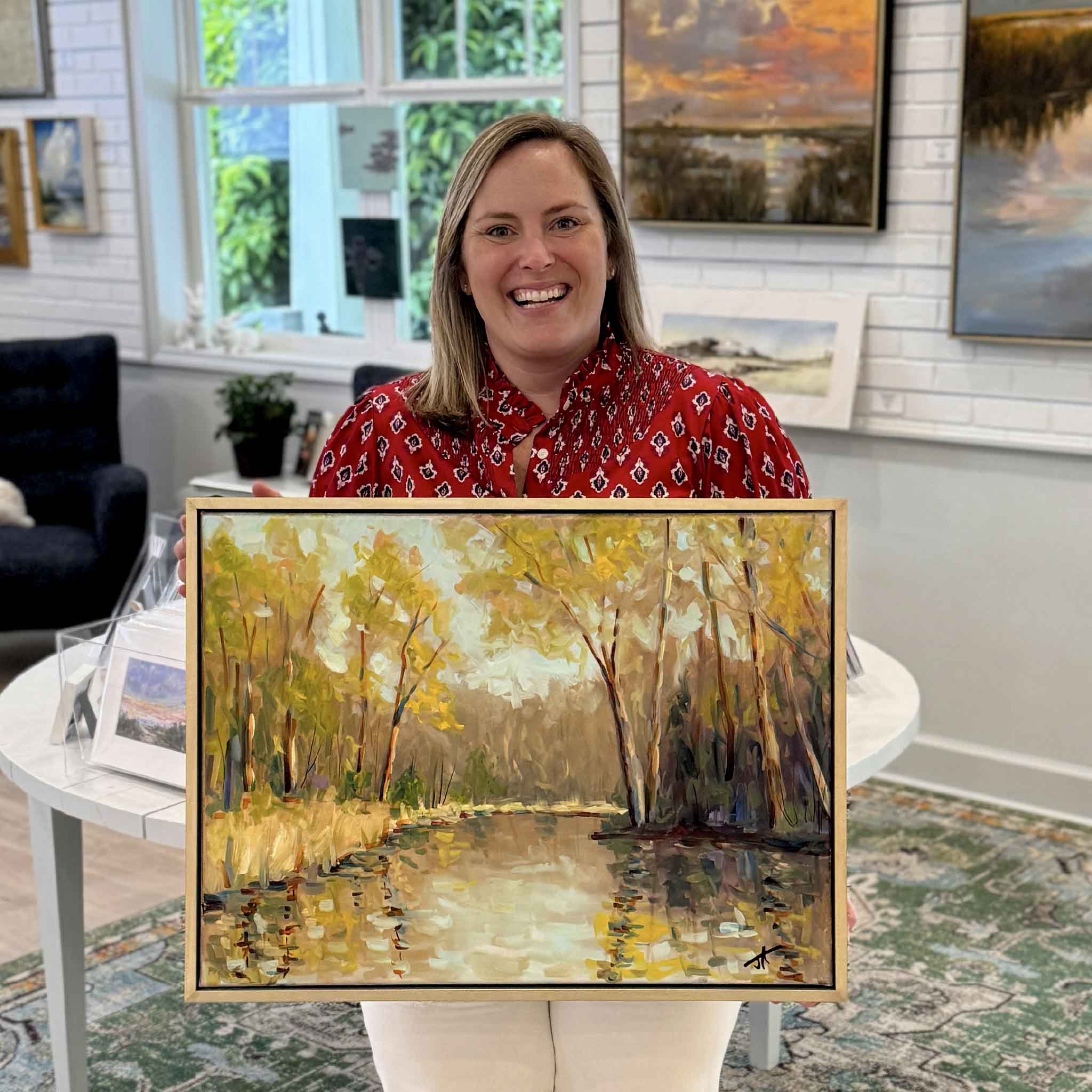 Bordy holding Spring Light by Jamie Arnold inside Gray Man Gallery in Pawleys Island, South Carolina, showing the scale of the framed original oil painting with reflective water, soft spring foliage, and luminous light, photographed in the gallery surrounded by coastal artwork and natural light