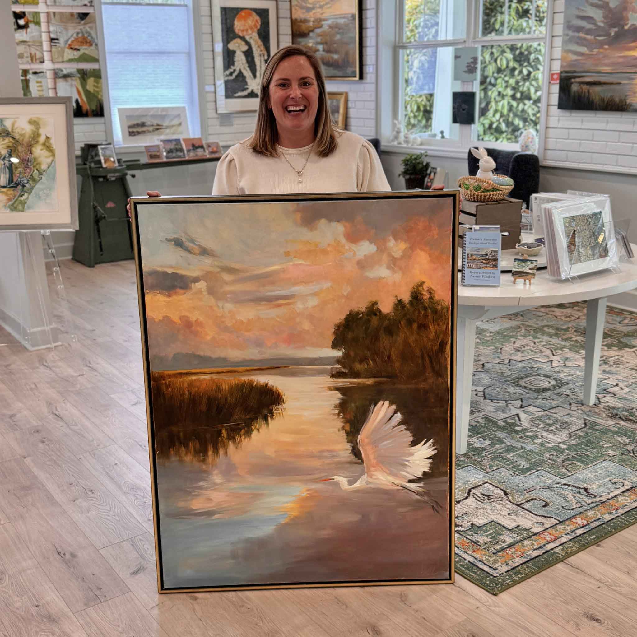 Bordy holding Sunset Cruise by local artist Helli Luck inside Gray Man Gallery Pawleys Island showing scale of large framed Lowcountry marsh painting with a white egret taking flight across glowing sunset water
