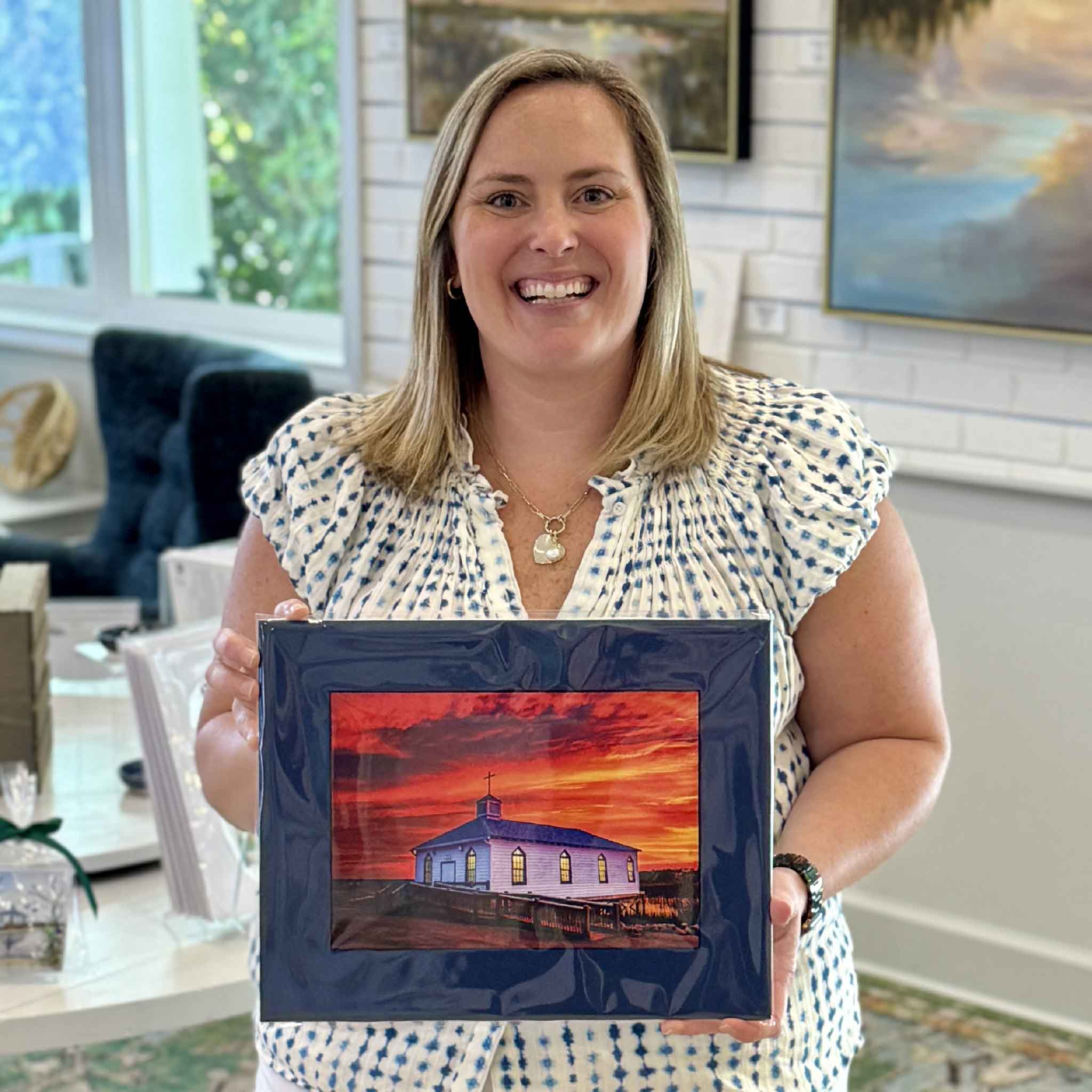 Bordy holding a matted Tanya Ackerman photography print at Gray Man Gallery in Pawleys Island, South Carolina. The artwork features the historic Pawleys Island chapel glowing beneath a dramatic Lowcountry sunset and was created from the cover of Tanya Ackerman’s book Pawleys Island: Seasons of Light.
