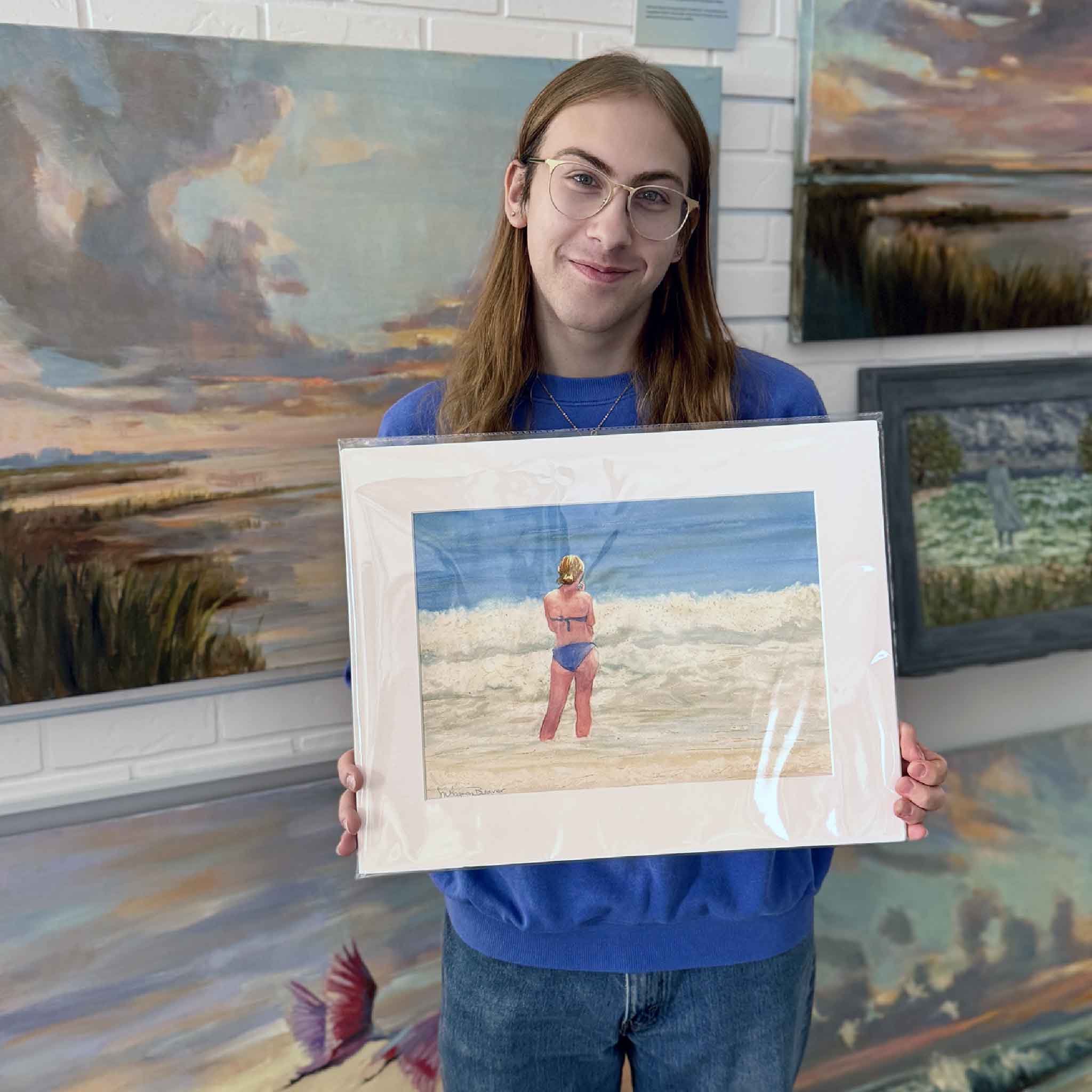 A Gray Man Gallery team member holds Can You Hear Me Now by Mimi Beaver, an original watercolor depicting a solitary figure standing at the ocean’s edge, donated to benefit Salty Dogs Senior Animal Rescue.