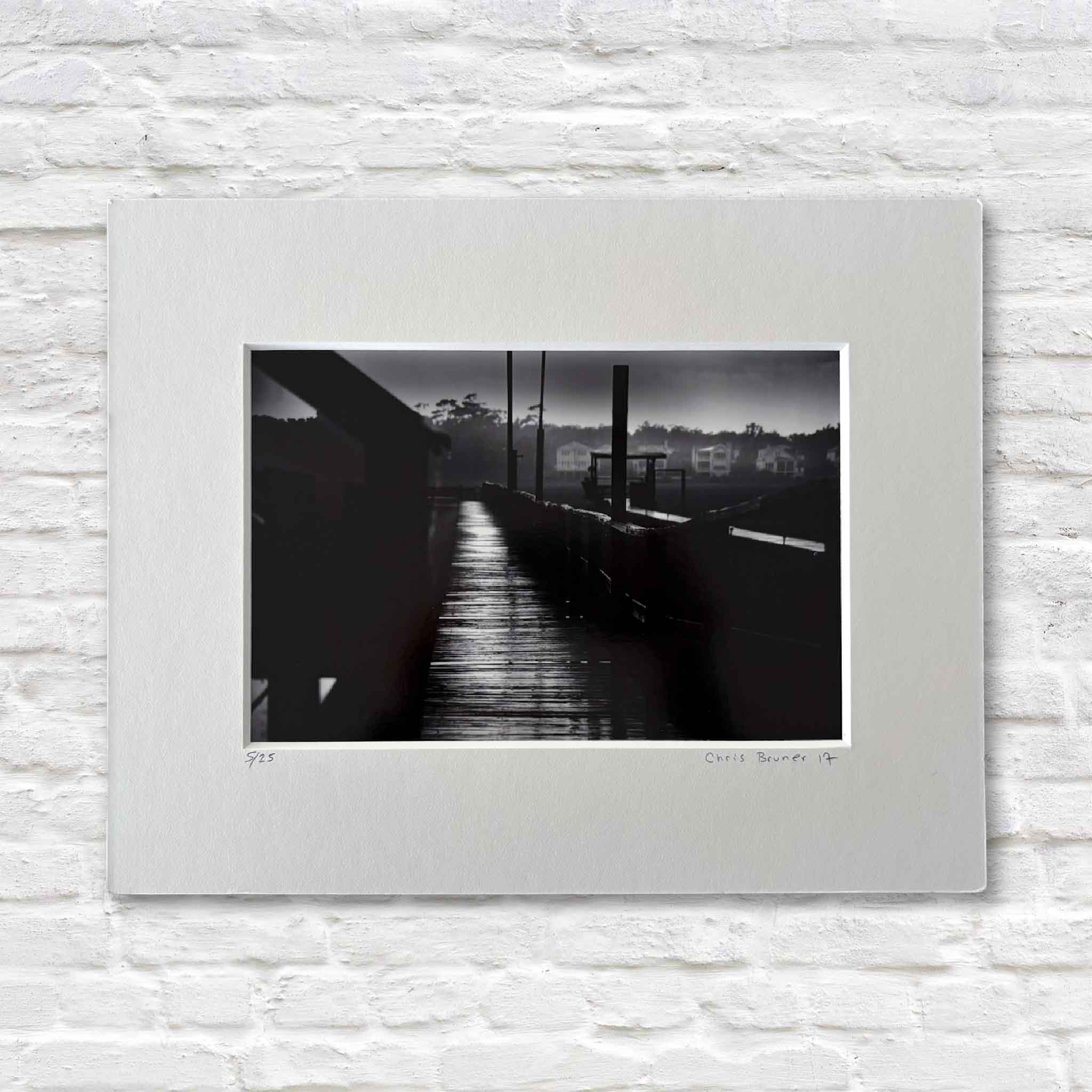Carolina #101, a signed and numbered black-and-white fine art photograph by local coastal photographer Chris Bruner, taken on Pawleys Island, South Carolina, featuring a quiet Lowcountry pier at dusk with reflective light, deep shadows, and calm coastal atmosphere, professionally matted and ready for framing.