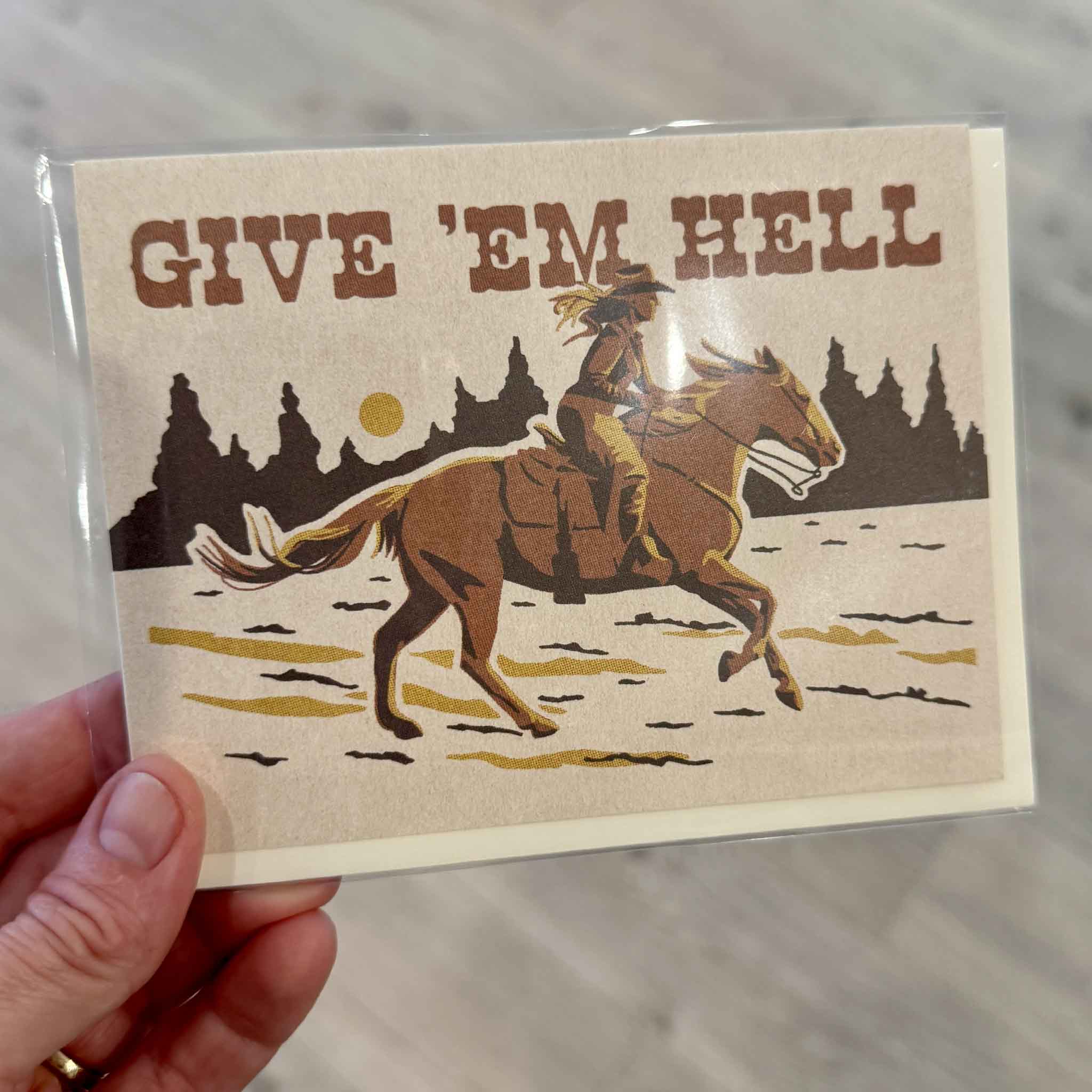 Give 'Em Hell encouragement greeting card illustrated by Caroline Clark for Red Cap Cards, photographed inside Gray Man Gallery in Pawleys Island, South Carolina. This blank-inside card features a Western cowgirl riding horseback across an open landscape with vintage typography and an inspiring message.