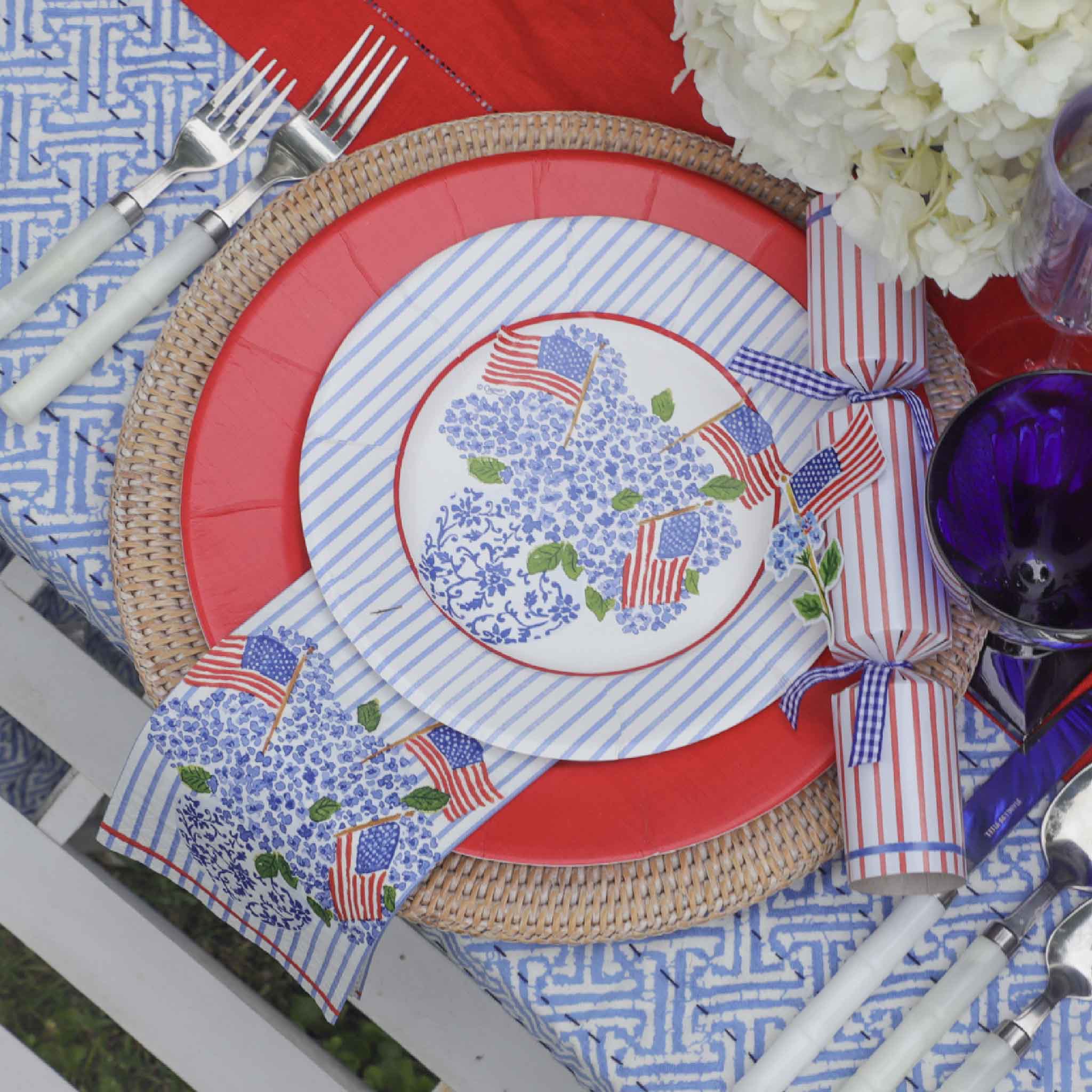 Caspari Americana Flags & Hydrangeas disposable paper plates and coordinating cocktail napkins by artist Lindsay Brackeen styled on a patriotic summer tablescape with woven chargers, bright red dinner plates, blue striped salad plates, white hydrangeas, American flag accents, and blue glassware. This premium entertaining collection is available at Gray Man Gallery in Pawleys Island, South Carolina, and is perfect for Fourth of July celebrations, Memorial Day gatherings, beach house entertaining, and summer 