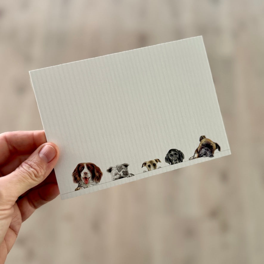 Front of Caspari Doggies flat correspondence card featuring Bob Bowdige artwork with five charming dogs peeking along the bottom edge, shown by hand at Gray Man Gallery in Pawleys Island, South Carolina. This premium flat correspondence card is blank for personal notes and sold as part of a boxed set of 12 cards with coordinating envelopes.