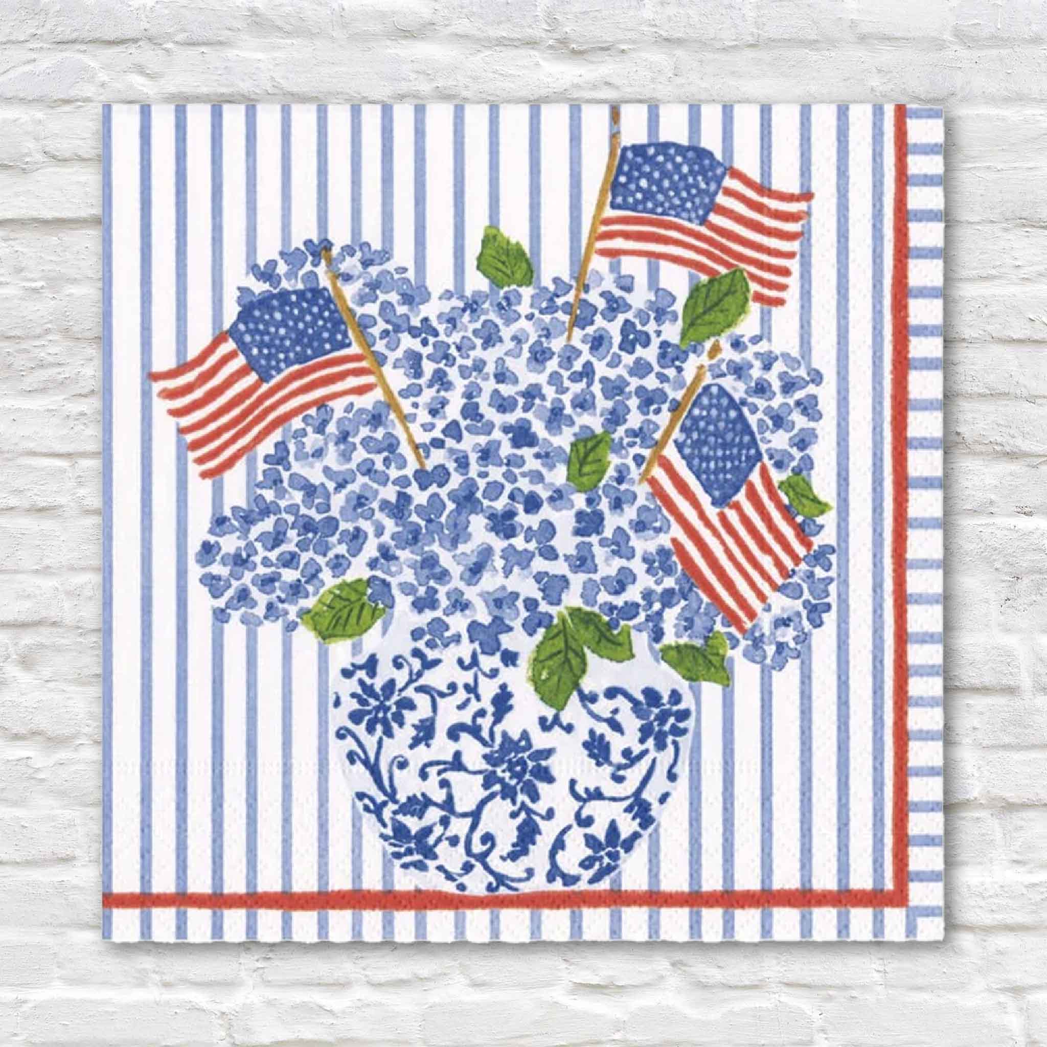 Caspari Flags & Hydrangeas cocktail napkin featuring artwork by Lindsay Brackeen, showcasing blue hydrangeas arranged in a blue-and-white chinoiserie vase accented with American flags on a blue striped background with a red border. Available at Gray Man Gallery in Pawleys Island, South Carolina. Each package includes 20 premium triple-ply paper cocktail napkins perfect for Fourth of July celebrations, Memorial Day, Labor Day, patriotic entertaining, coastal homes, and elegant gatherings.