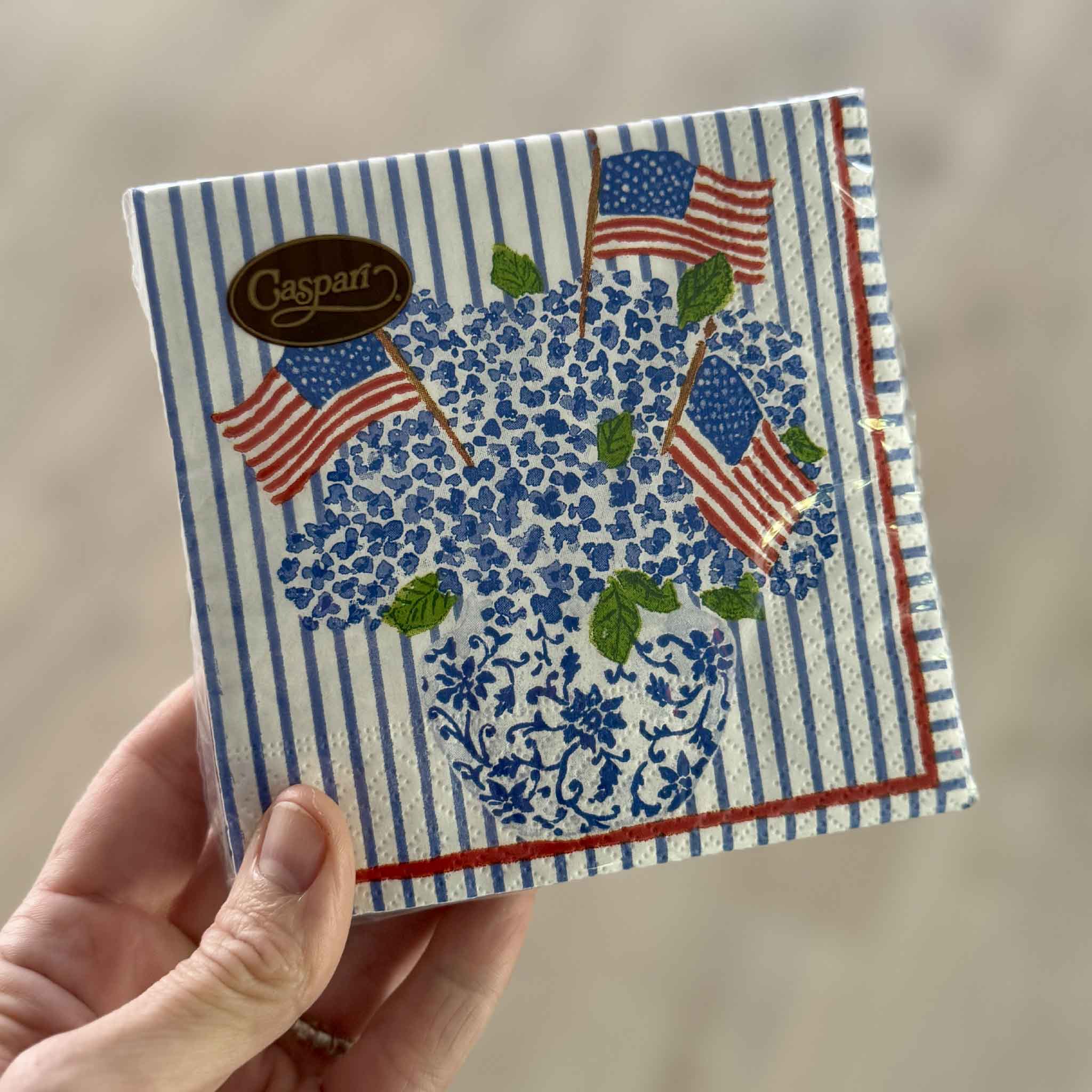Retail package of Caspari Flags & Hydrangeas cocktail napkins by artist Lindsay Brackeen displayed at Gray Man Gallery in Pawleys Island, South Carolina. Each package contains 20 premium triple-ply 5 x 5 inch paper cocktail napkins featuring patriotic blue hydrangeas in a chinoiserie vase, ideal for Fourth of July parties, Memorial Day gatherings, Labor Day celebrations, and elegant entertaining.