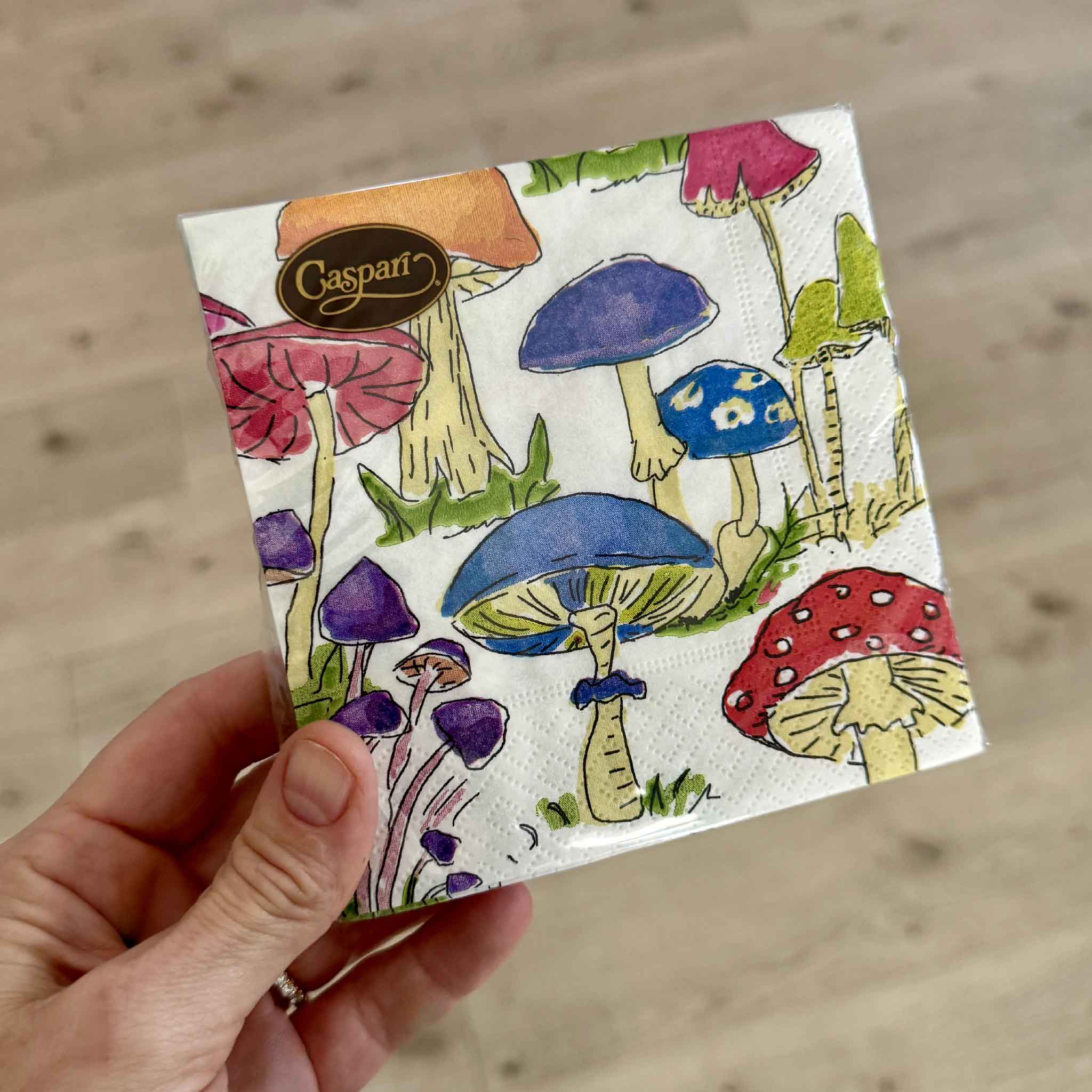 A hand holds a package of Caspari Woodland Mushrooms cocktail napkins inside Gray Man Gallery in Pawleys Island, South Carolina. Featuring vibrant watercolor illustrations of colorful mushrooms, these FSC-certified triple-ply napkins are printed in Germany with non-toxic, water-based inks and make a charming hostess gift for mushroom lovers, gardeners, nature enthusiasts, and cottagecore décor fans.