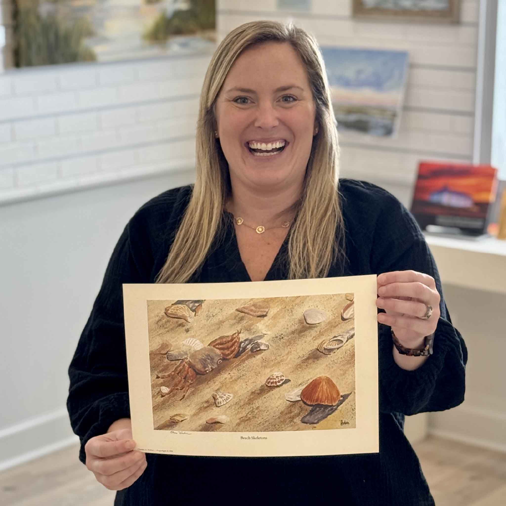 Gray Man Gallery staff member Bordy holding Beach Skeletons print by Pawleys Island artist Charles Huber to show scale, featuring crab shell and coastal beach treasures rendered in soft realism.