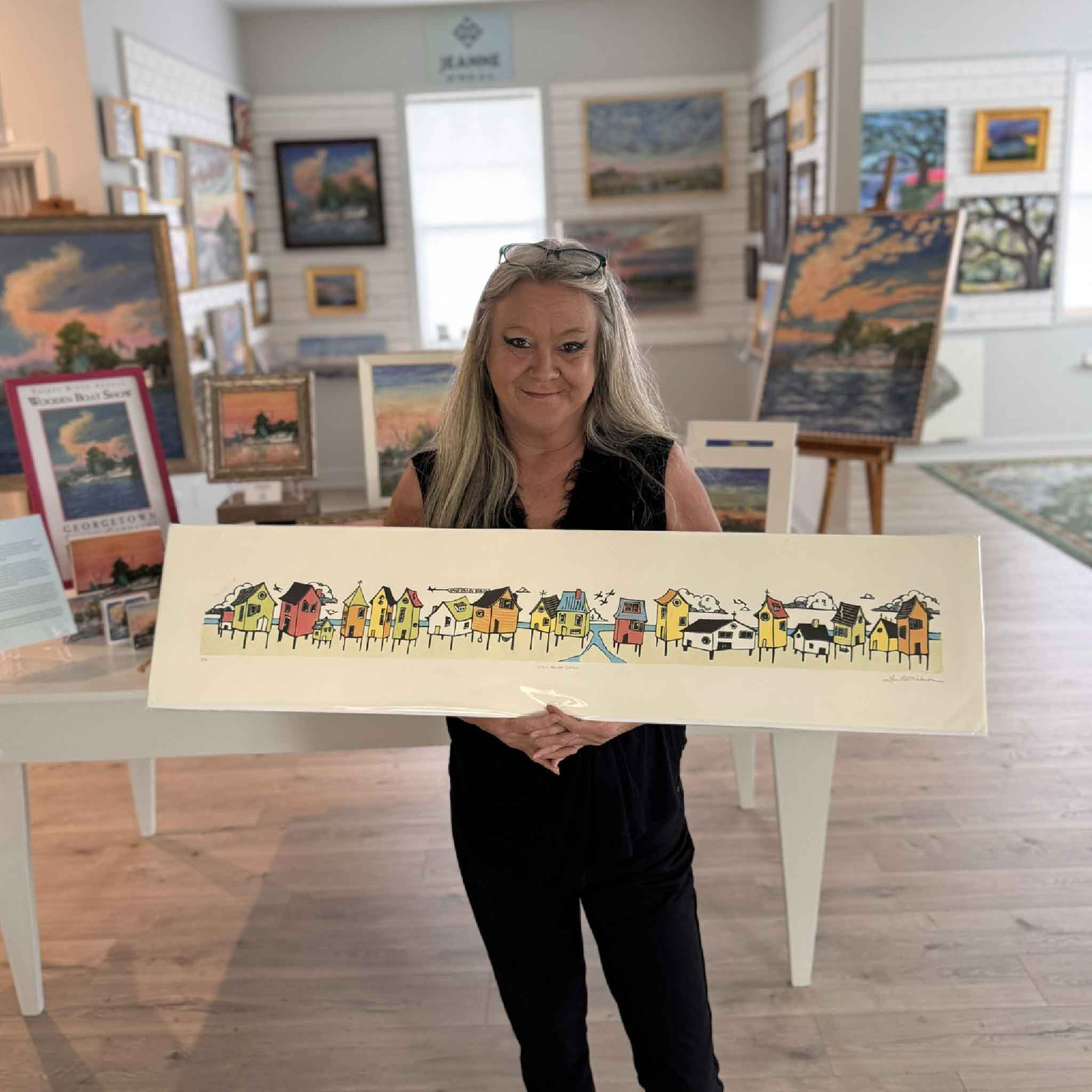 Charlotte holding Laurie McIntosh's original Stick House Beach linocut at Gray Man Gallery. The colorful coastal artwork features a lively row of whimsical beach cottages rendered in Laurie's signature hand-pulled linocut reduction style.