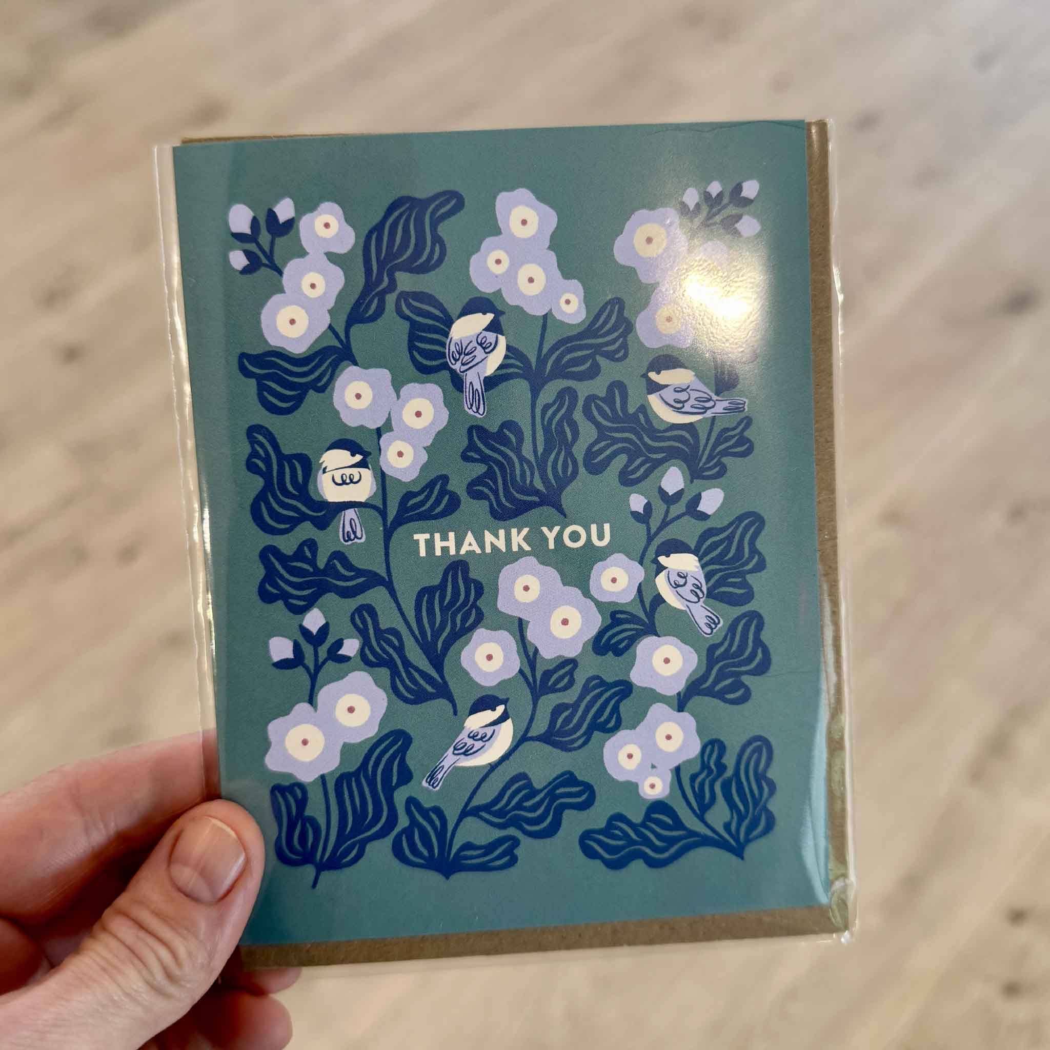 Customer holding the Chickadee Thank You Greeting Card by Nuthatch Studio inside Gray Man Gallery in Pawleys Island, South Carolina. This charming greeting card features delicate white blossoms and illustrated chickadees perched among leafy branches on a soft teal background with a "Thank You" message, printed on premium 100% recycled paper and paired with a recycled kraft envelope.