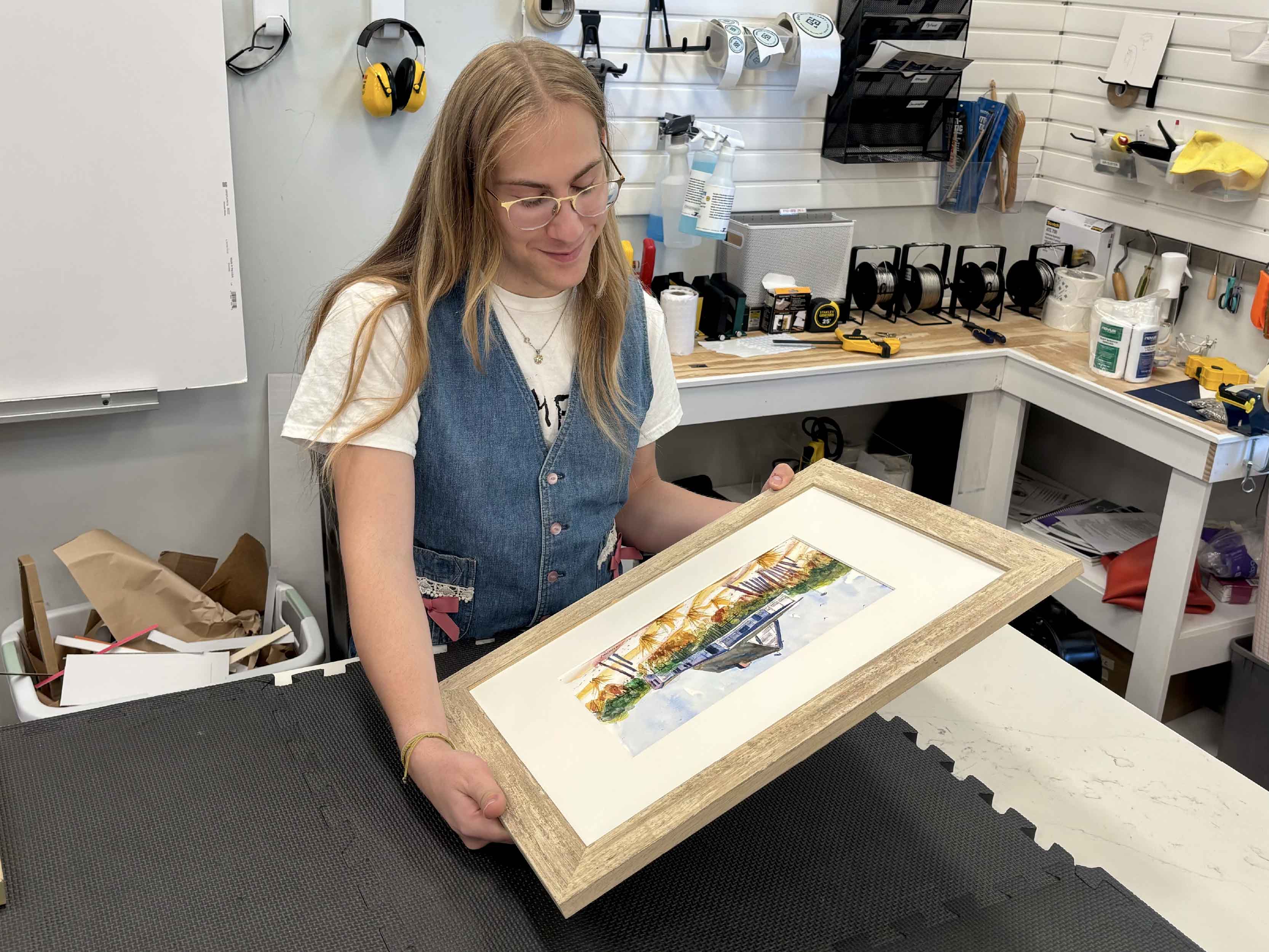 Gray Man Gallery team member carefully inspects a completed custom-framed watercolor in the professional frame shop at Gray Man Gallery in Pawleys Island, South Carolina. The finished artwork is shown with custom matting and a coordinating wood frame during the final quality inspection, when the frame, corners, glazing, mat, artwork, and finishing details are checked before the completed custom framing project is returned to the customer.