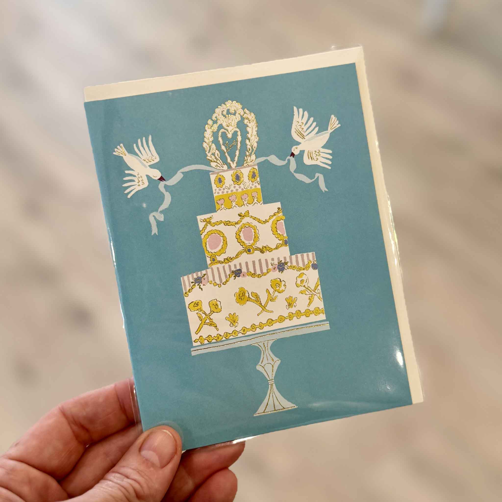 Love Cake wedding greeting card illustrated by Danielle Kroll for Red Cap Cards, photographed inside Gray Man Gallery in Pawleys Island, South Carolina. This elegant blank-inside greeting card features a beautifully illustrated tiered wedding cake with shimmering gold foil accents, white doves carrying blue ribbons, and a romantic heart topper, making it perfect for weddings, engagements, anniversaries, and bridal showers.