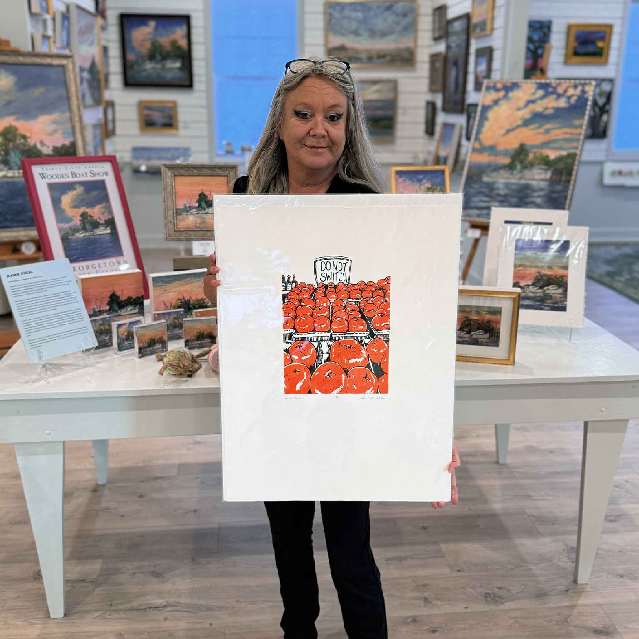 Charlotte holding “Do Not Switch,” an original linocut by Laurie McIntosh featuring rows of bright orange tomatoes with a playful sign.