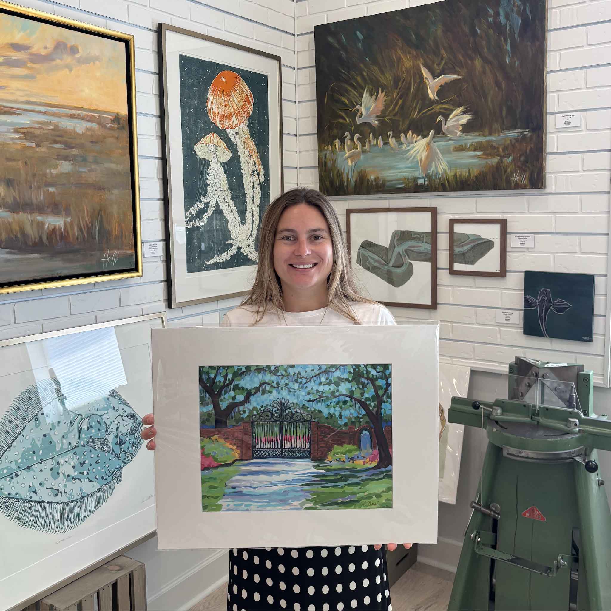 Kyra holding a larger 16x20 professionally matted Litchfield Gate fine art print by South Carolina artist Elizabeth Sheats inside Gray Man Gallery in Pawleys Island, featuring the historic Litchfield Plantation gates, live oaks, Spanish moss and colorful Lowcountry landscaping.