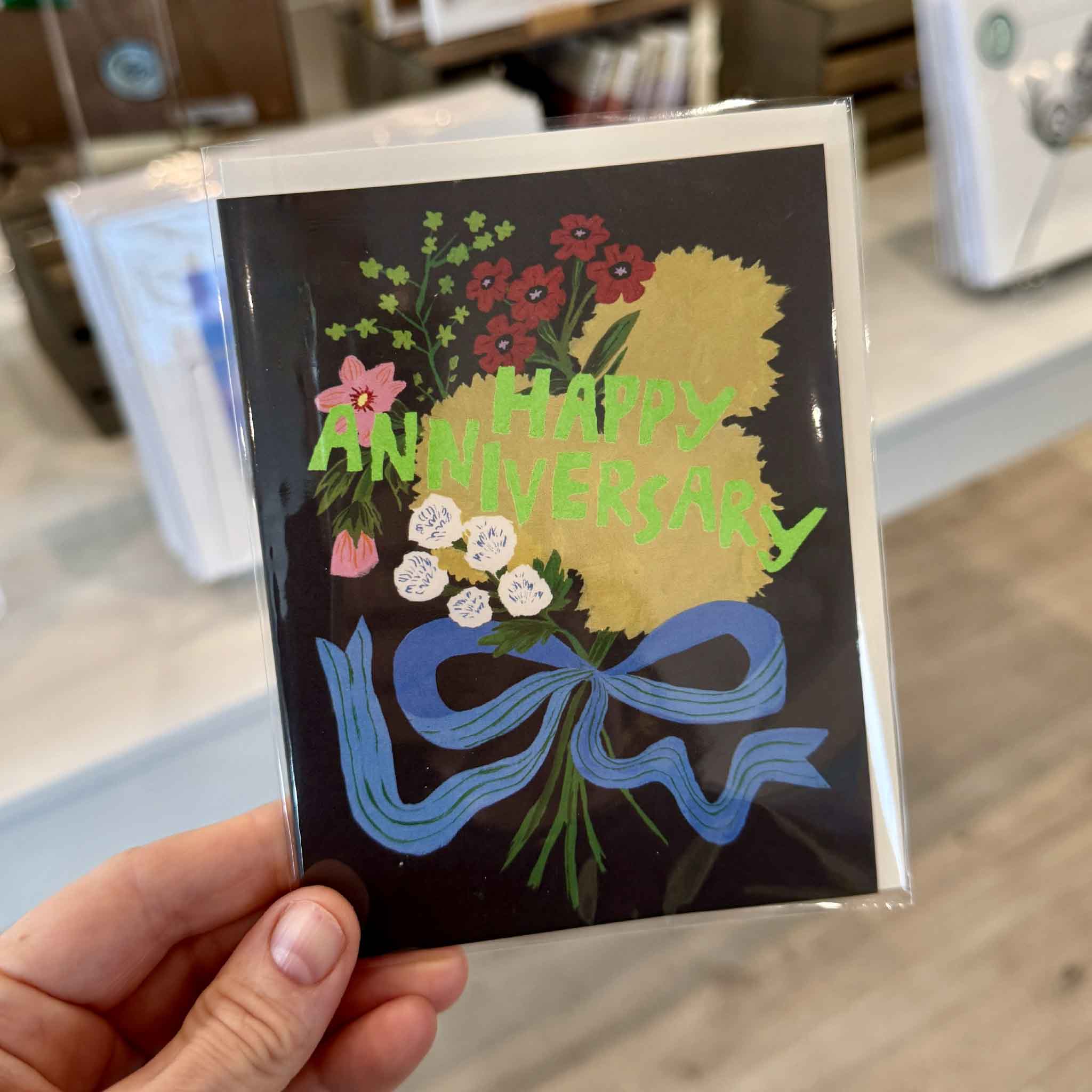 Emily Isabella Anniversary Bouquet greeting card held inside Gray Man Gallery in Pawleys Island, featuring a whimsical floral arrangement with colorful blooms, bright green Happy Anniversary lettering and a flowing blue bow on a dark background.
