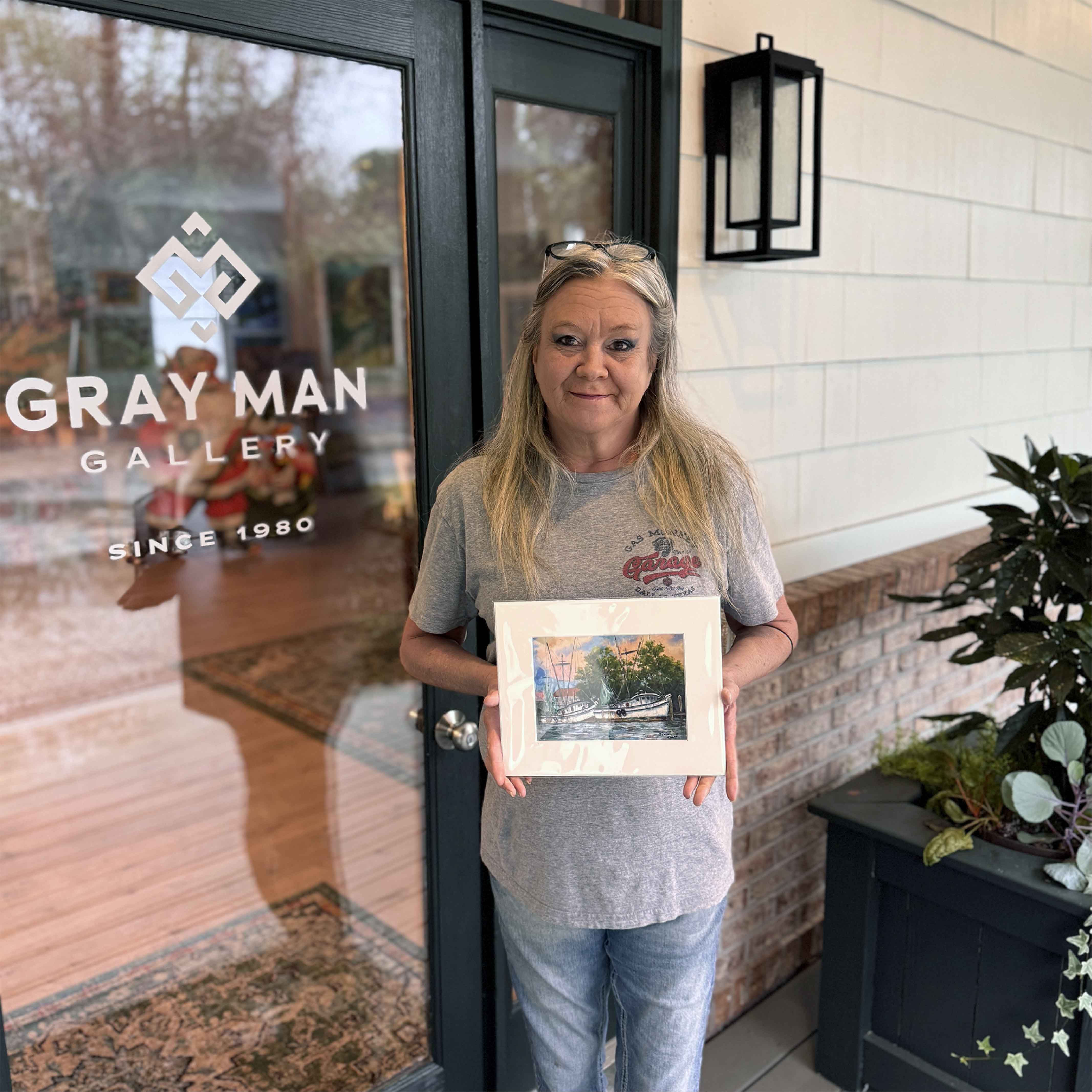 Charlotte holding fine art print of End of Day by Jeanne O’Neal showing Georgetown shrimp boats at sunset, matted and ready to frame. Pictured at Gray Man Gallery.