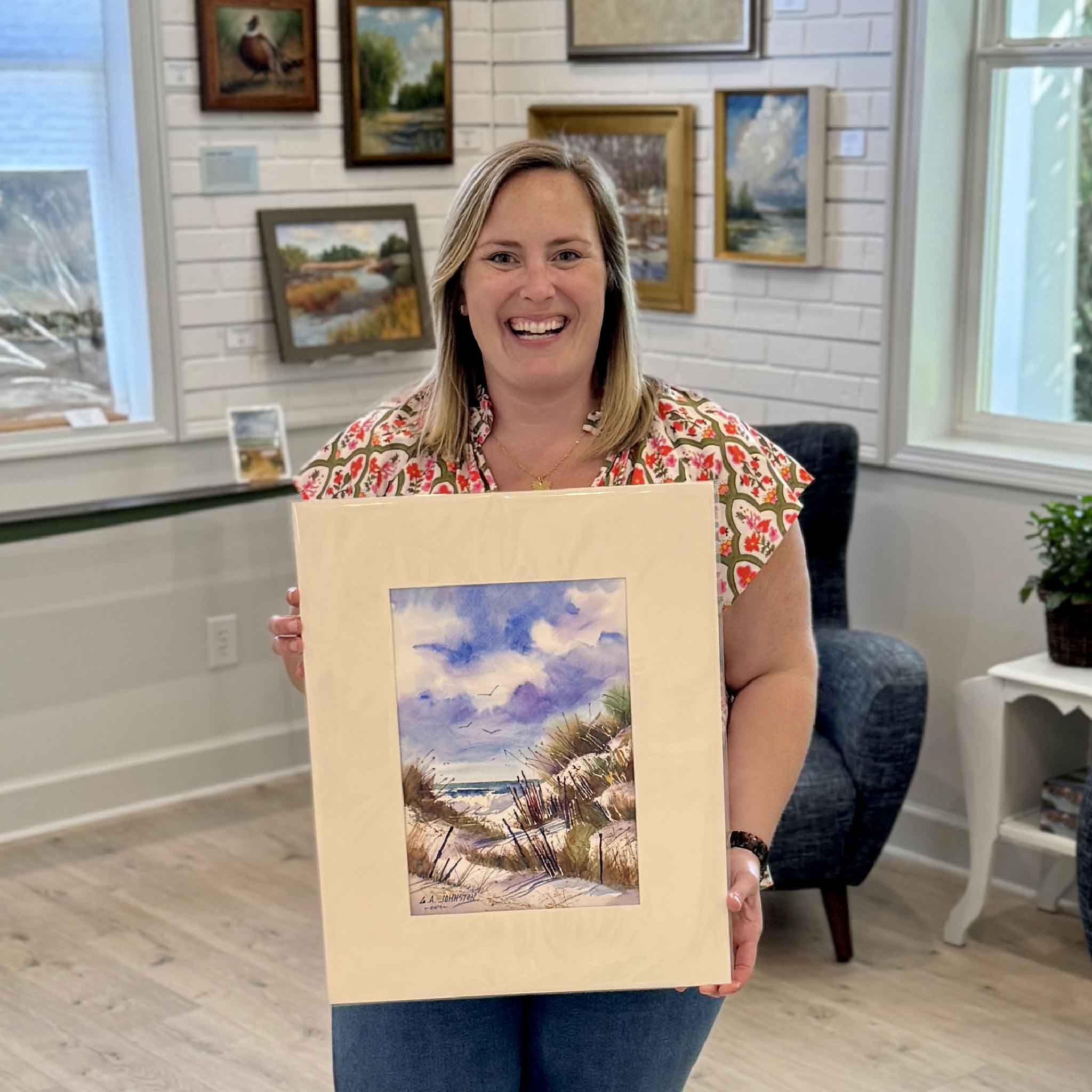 Bordy holding an original Gary Johnston watercolor titled Dunes at First Light inside Gray Man Gallery in Pawleys Island, showing scale and presentation of the artwork featuring coastal dunes, fencing, and distant ocean light, professionally matted to fit a standard 16x20 frame and ready for custom framing or a ready-made frame